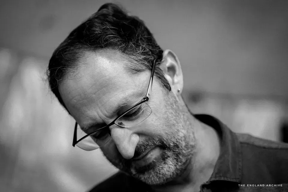 Close portrait of Lewis from above, head bowed at the work, the curve of his glasses and the small grey hairs in his beard caught in the diffuse light from the canopy.