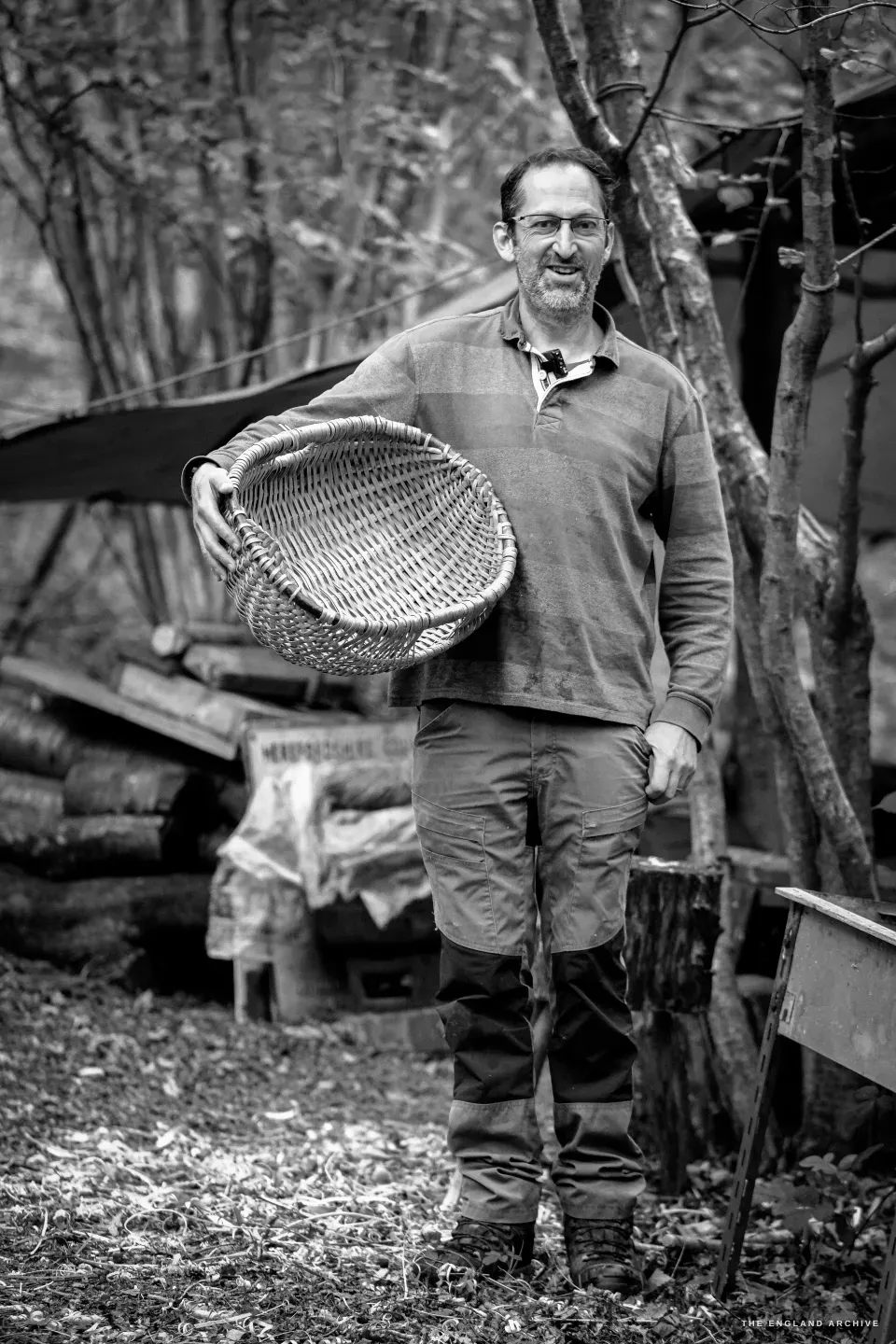 Lewis Goldwater standing in front of the woodland workshop holding a freshly finished Whisket basket in his left hand, smiling at the camera, the canopy and the rod-pile of his working life behind him.