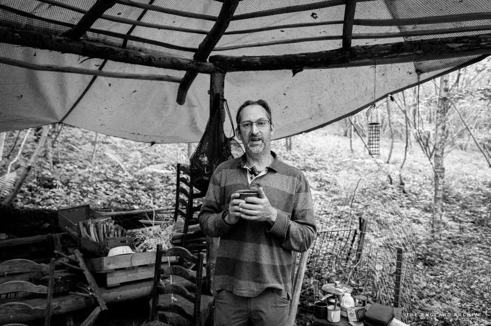 Lewis under the translucent plastic canopy of his workshop, holding a mug of tea in both hands, looking calmly at the camera. Stacked baskets and rod bundles flank him.