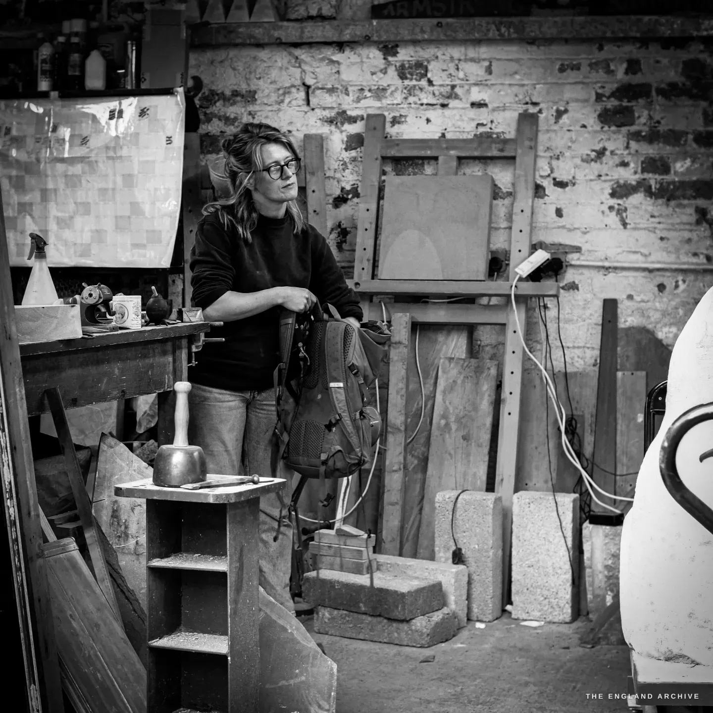 Lily Marsh standing by her bench with a rucksack in her hands, looking off to one side, a dummy mallet and a chisel resting on a shelf in the foreground, the crowded workshop behind her.