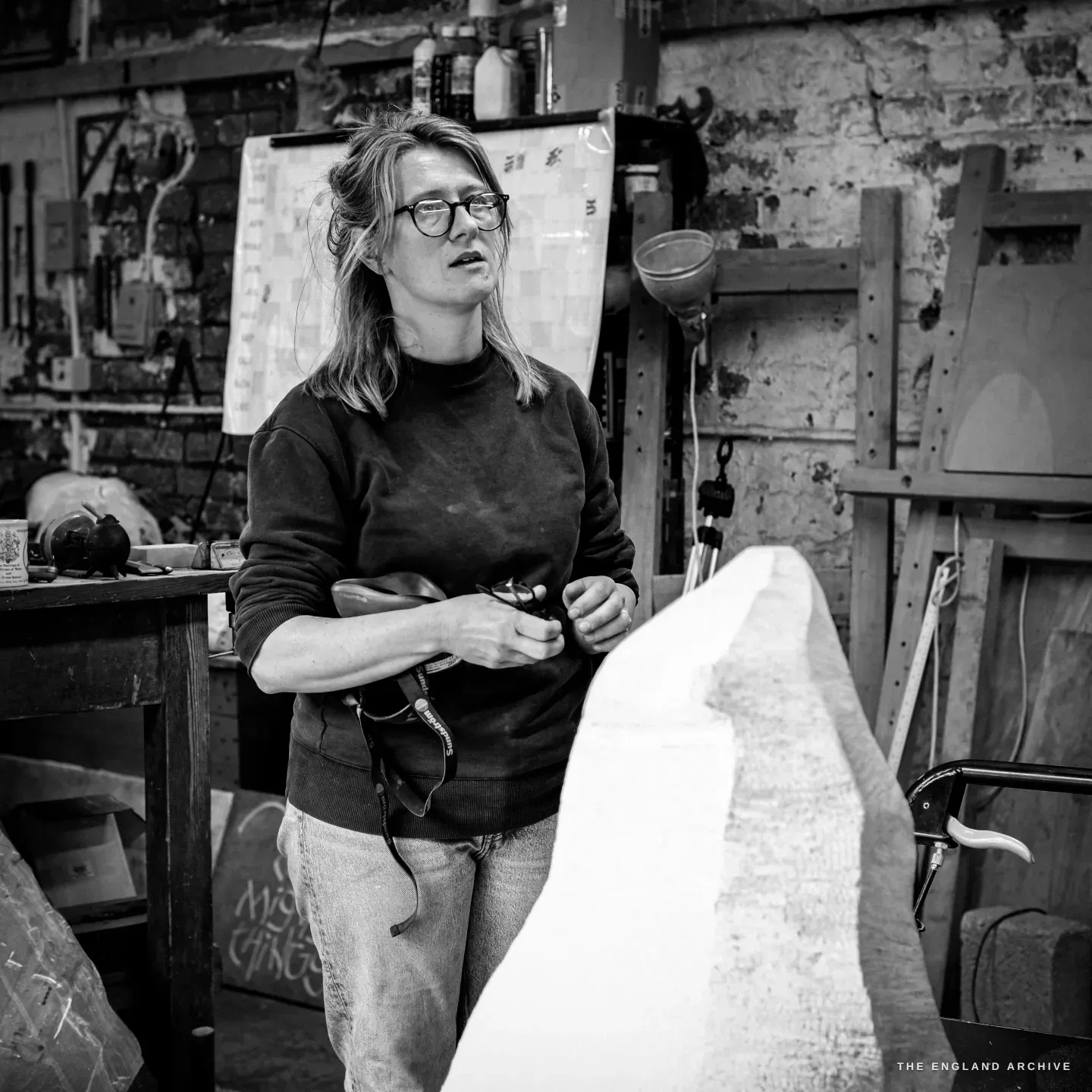 Lily Marsh mid-conversation, holding her glasses in one hand, the roughed-out white stone rising in the foreground and a large paper template pinned to the brick wall behind.