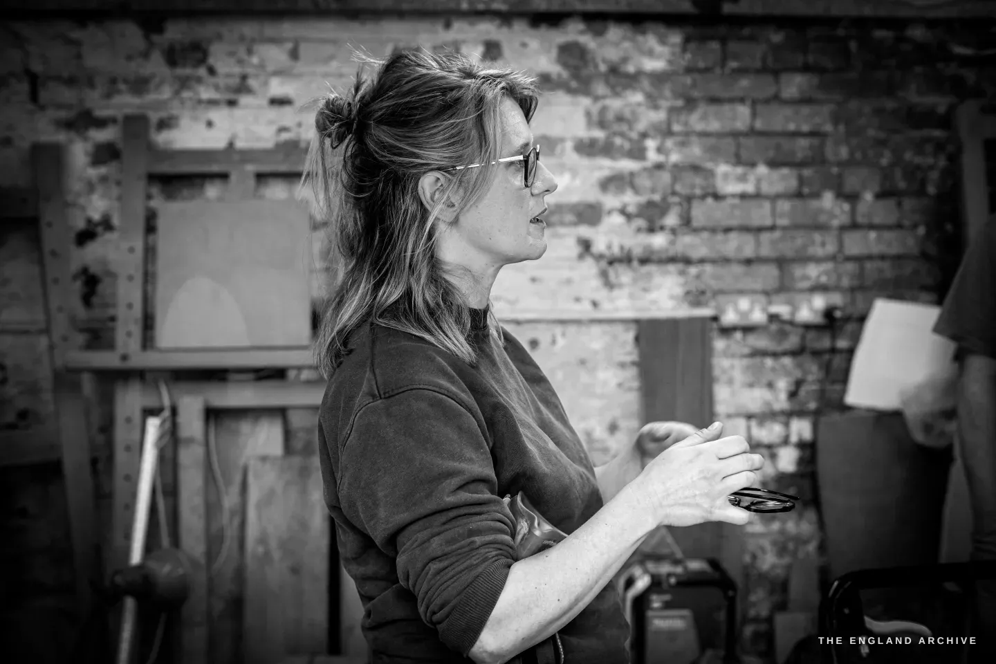 Lily Marsh in profile, hair tied back, talking with her hands raised, the bare brick wall of the workshop behind her and a maker working at the right edge of the frame.