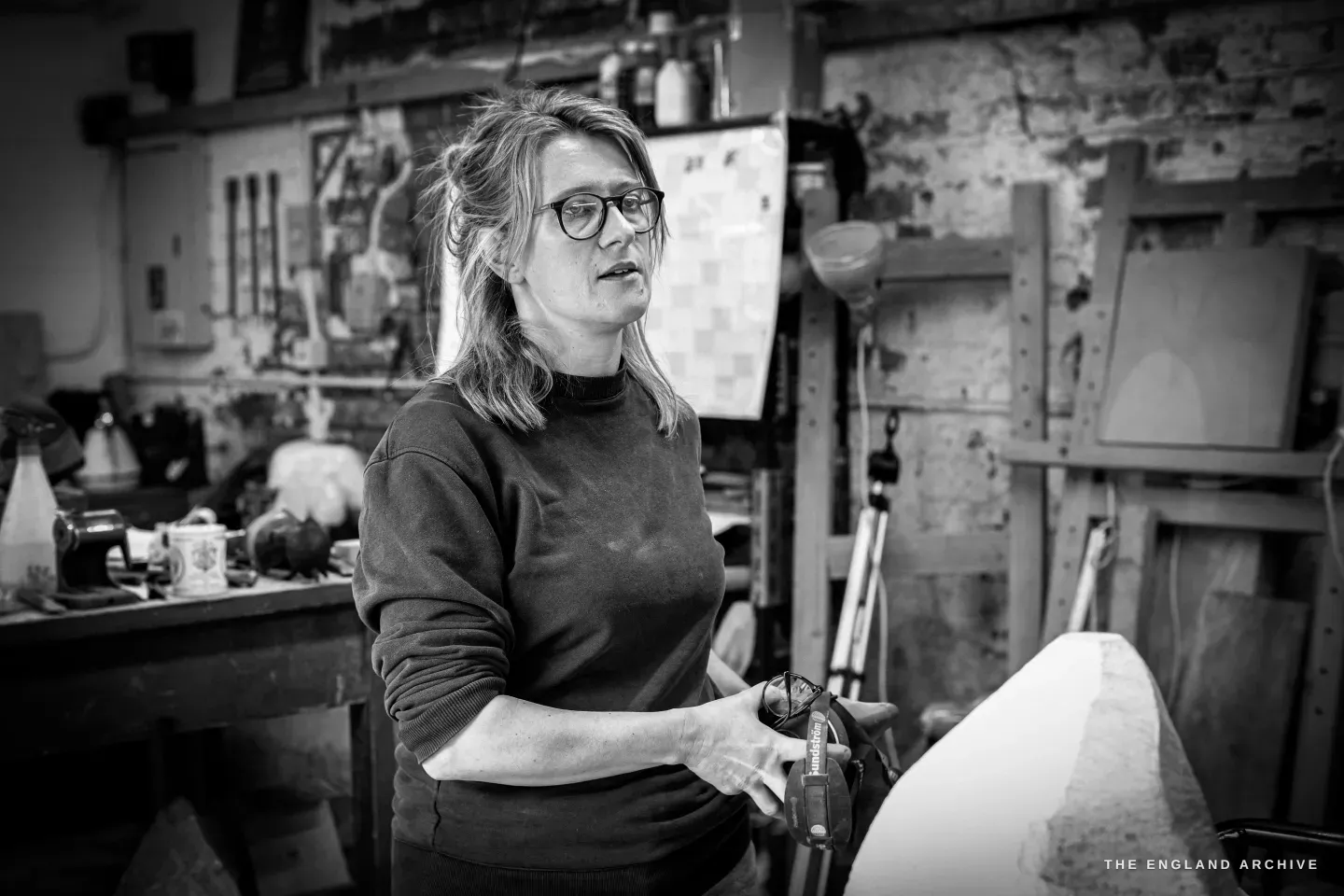 Lily Marsh mid-sentence holding a chisel loosely in both hands, the white sculpture catching the light at the right edge of the frame, shelves of tools and bottles behind.
