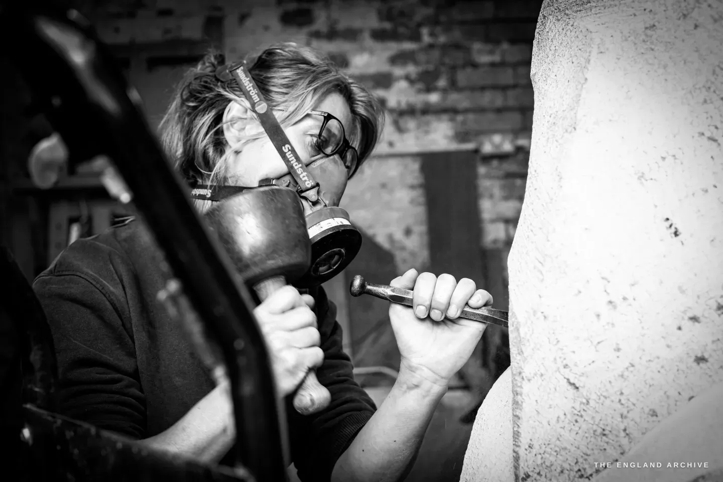 Close view of Lily Marsh carving, respirator over her mouth and glasses pushed up, both hands working a chisel against the stone, the wooden mallet blurred in motion.