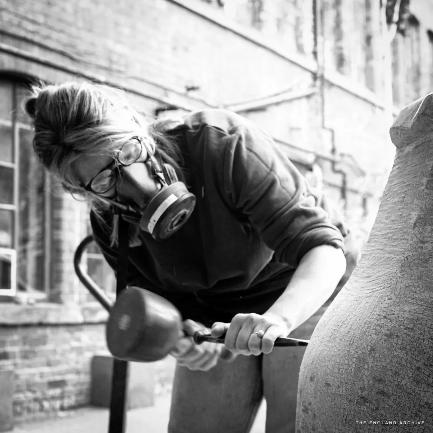 Lily Marsh carving out in the works yard, bent low over the stone in a respirator, mallet and chisel in her hands, the long brick range of Stag Works receding behind her.