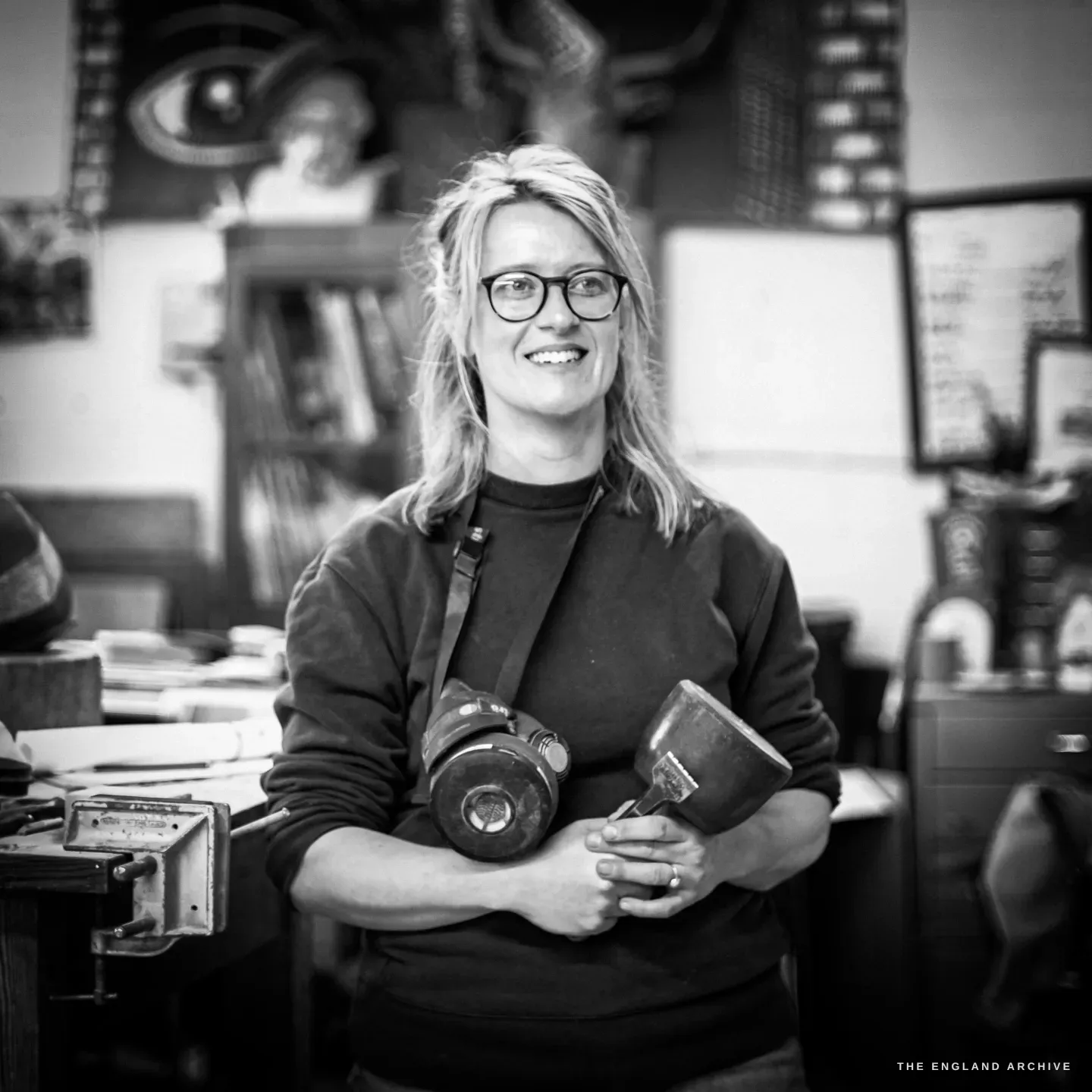 Lily Marsh smiling at the camera in her workshop, holding a respirator and a dummy mallet against her chest, glasses on, a large artwork of an eye and antlers on the wall behind her.