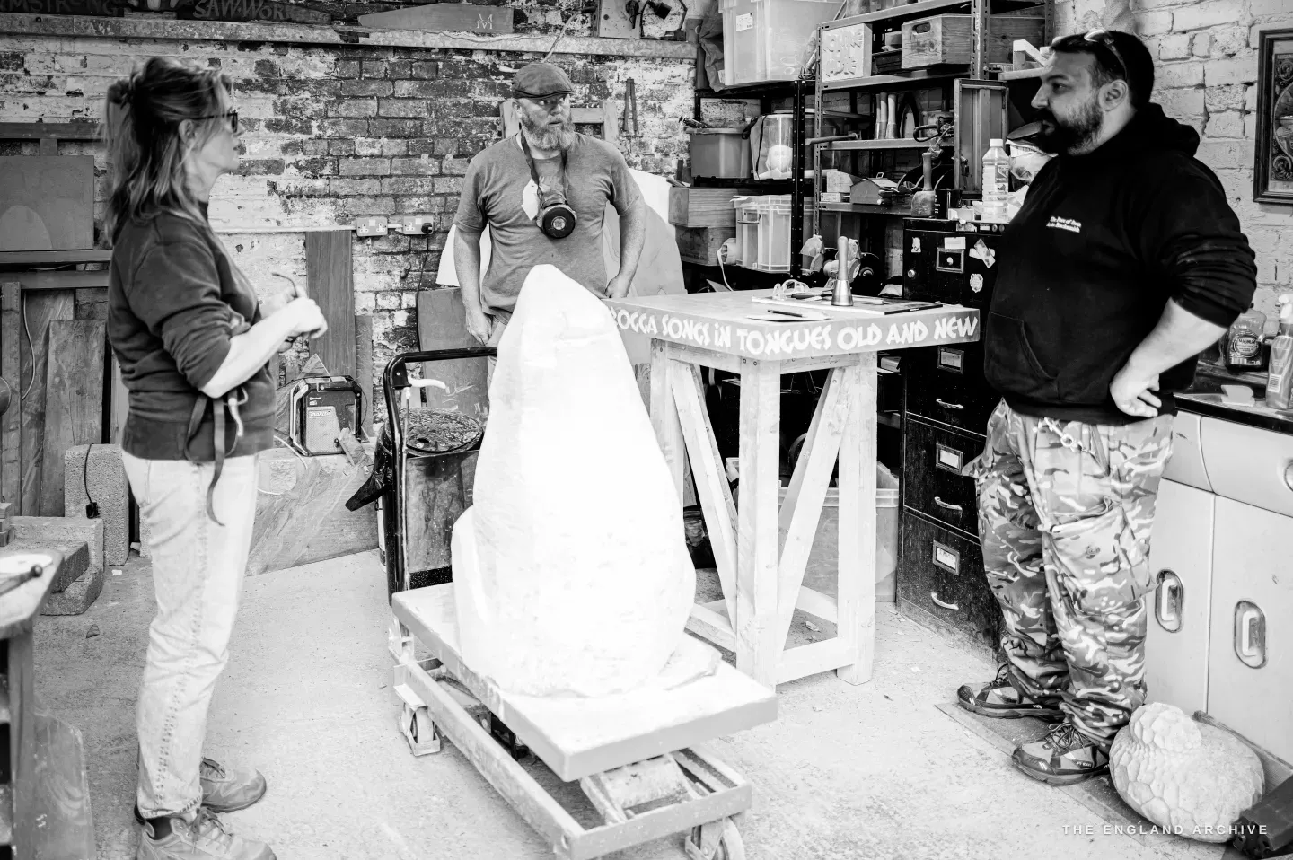 Three people standing inside the workshop around a tall block of white stone on a wheeled trolley - Lily Marsh at left in a work apron, a man in a cap with a respirator round his neck at centre, and a bearded visitor in a dark hooded top at right, brick walls and crowded shelving behind them.
