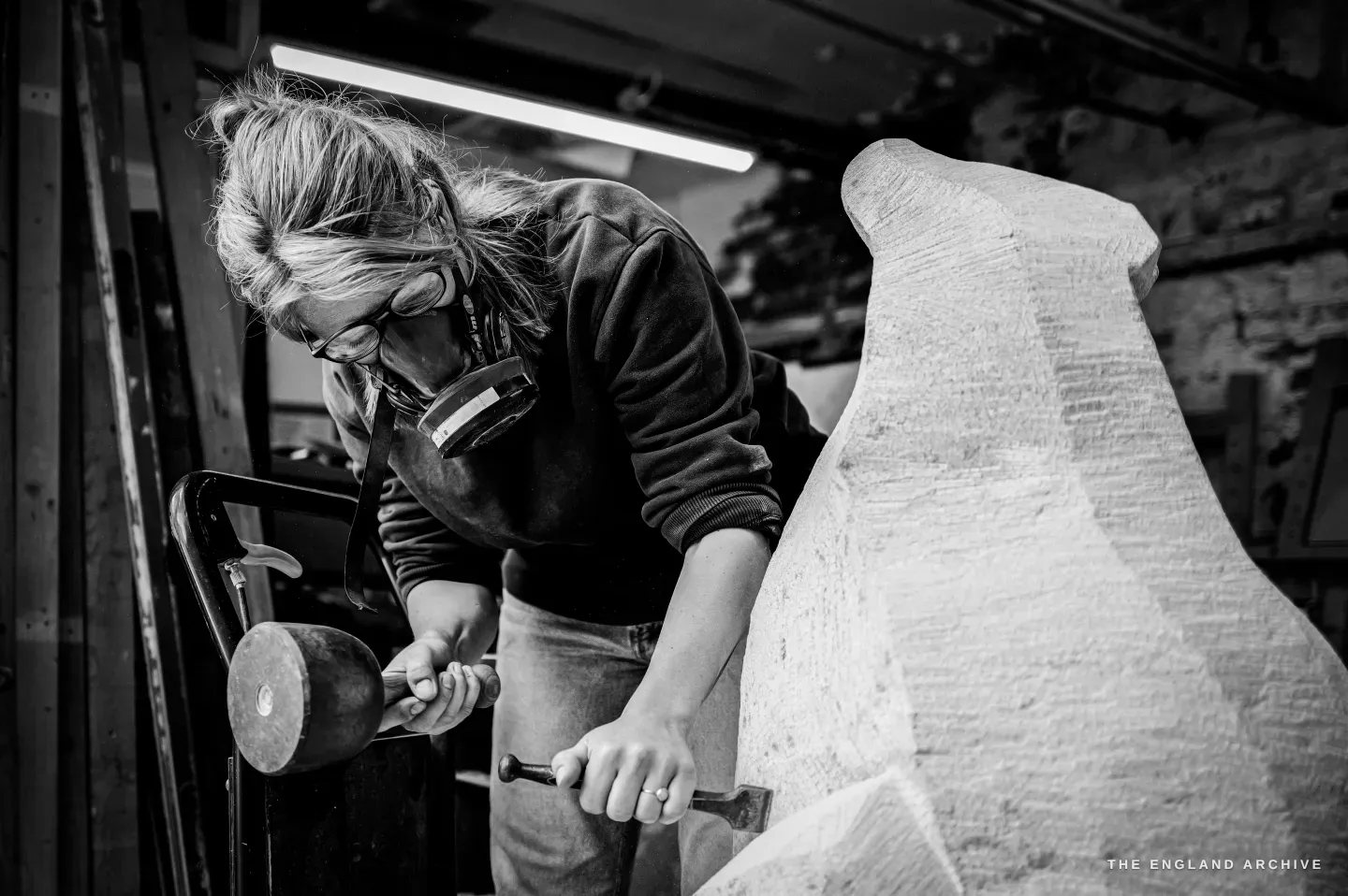 Lily Marsh in a respirator and glasses, leaning into the work, a round-headed dummy mallet raised in one hand and a chisel held to the pale stone in the other, the tall carved form rising on the right.