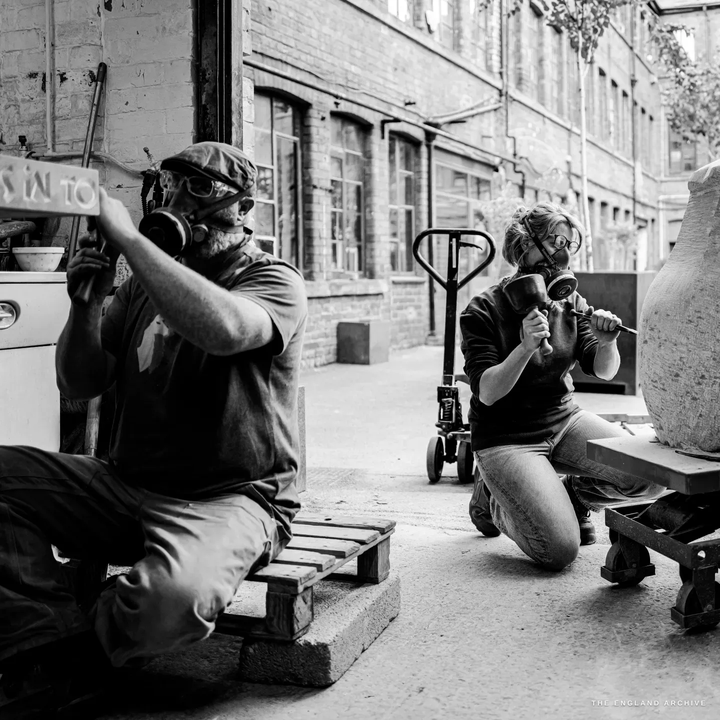 Steve and Lily Marsh working out in the yard at Stag Works - Steve at left cutting a stone sign that reads ‘songs in to…’, a respirator on his face, Lily at right kneeling and carving her standing stone, the long industrial range behind them.