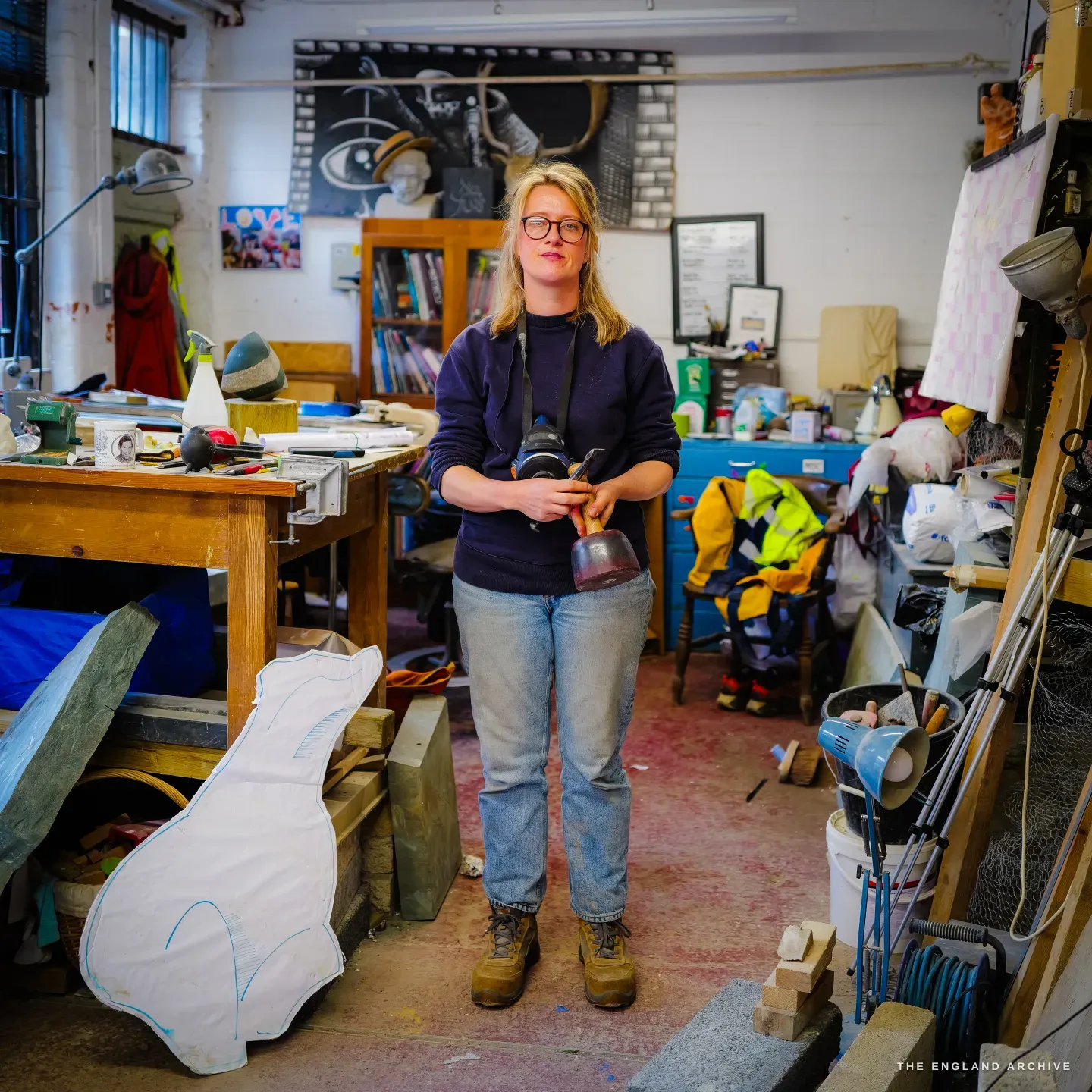 Lily Marsh standing in the middle of the shared workshop holding a dummy mallet and chisel, a large stone template with a blue drawn outline propped at her left, the crowded studio around her.