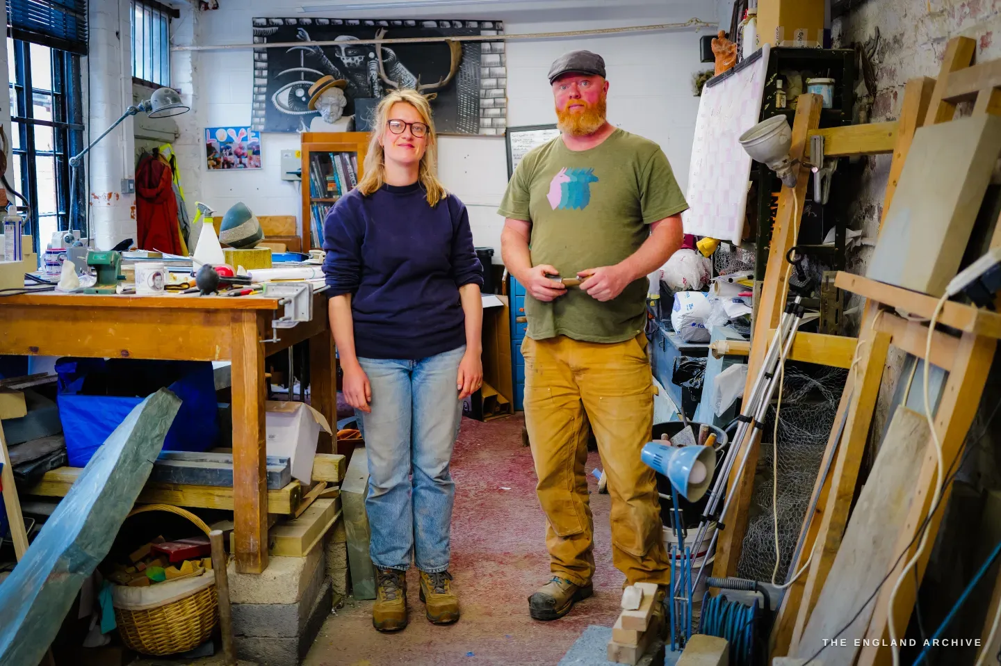 Lily Marsh and Steve standing together in the middle of the workshop, facing the camera - Lily at left in a dark jumper, Steve at right with a full beard and cap, the bench and racked stone slabs around them.