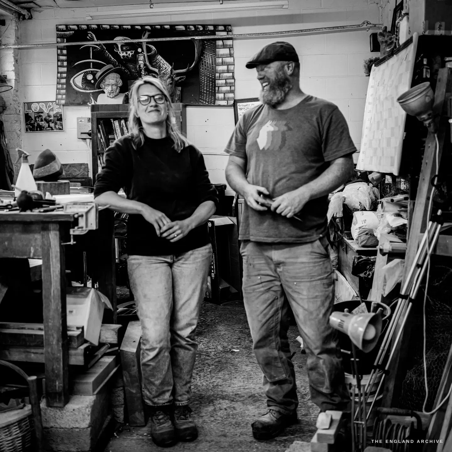Lily Marsh leaning against her bench with her arms crossed, smiling sideways, while Steve stands beside her laughing, a large dark artwork of an eye and antlers on the wall behind them.