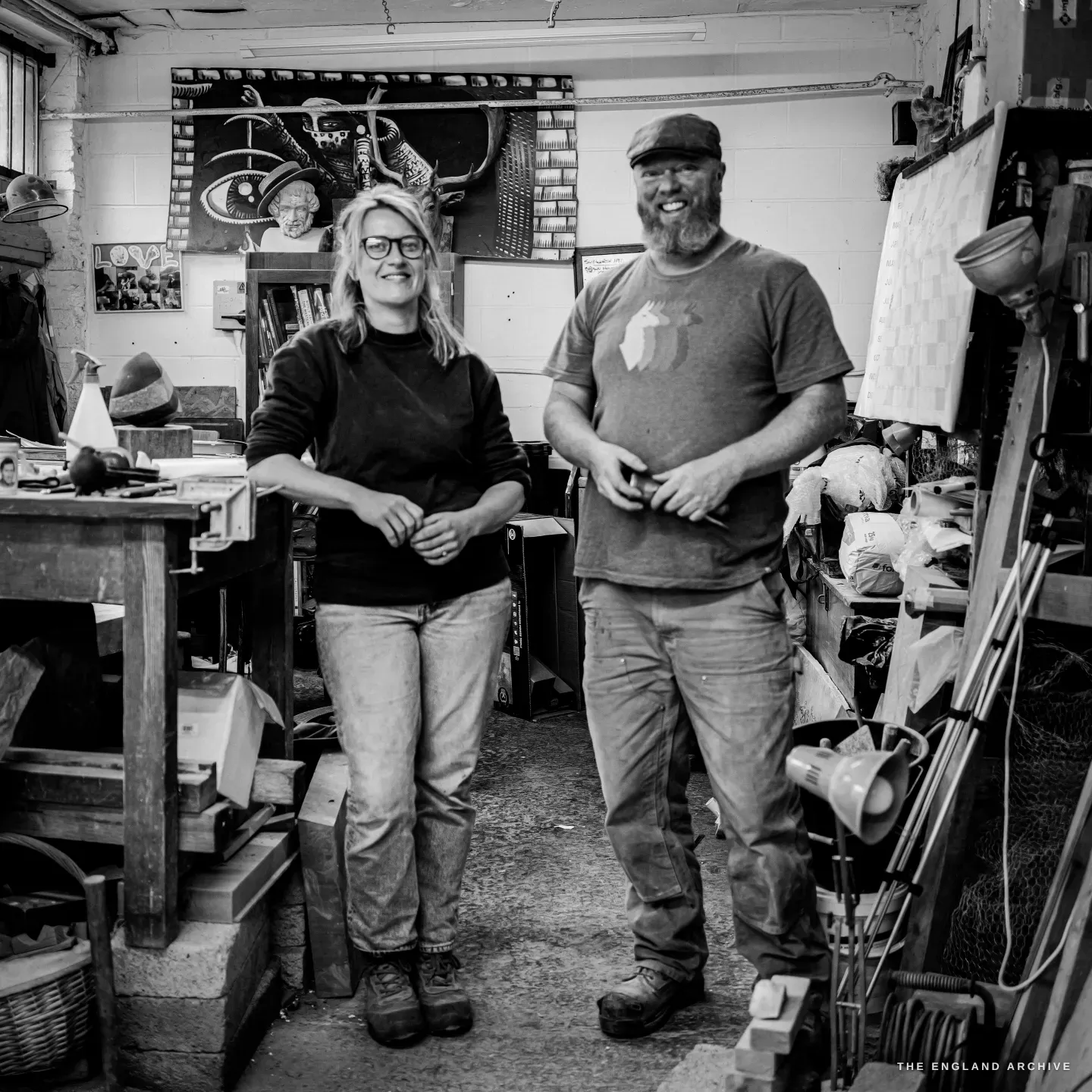 Lily Marsh and Steve standing side by side in the workshop, both turned to the camera and smiling, tools and shelving crowding the frame around them.