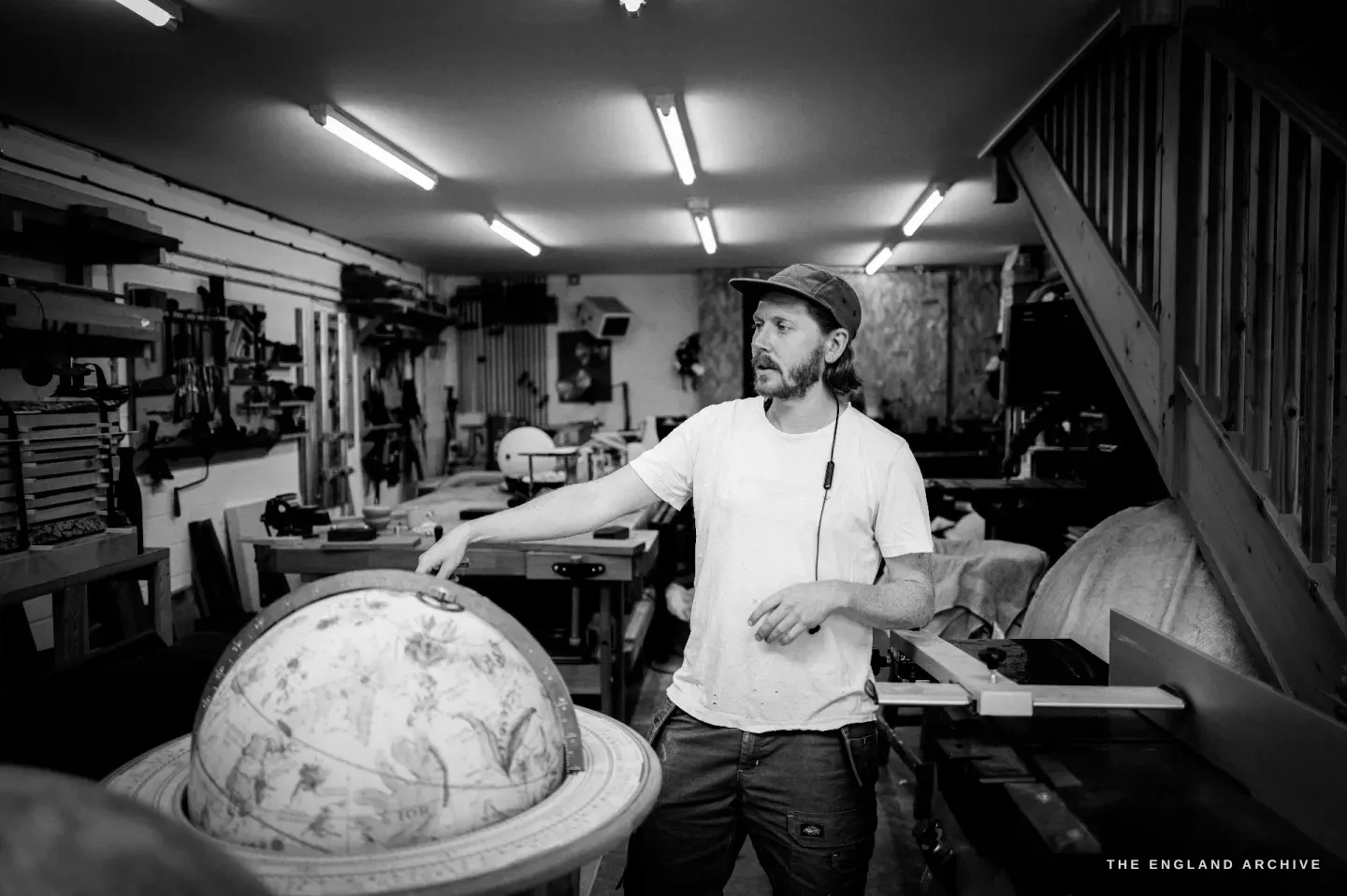 Louis Craig Carpenter standing in his ground-floor workshop, a hand on a large globe on its stand, the staircase up to the globemaker behind him.