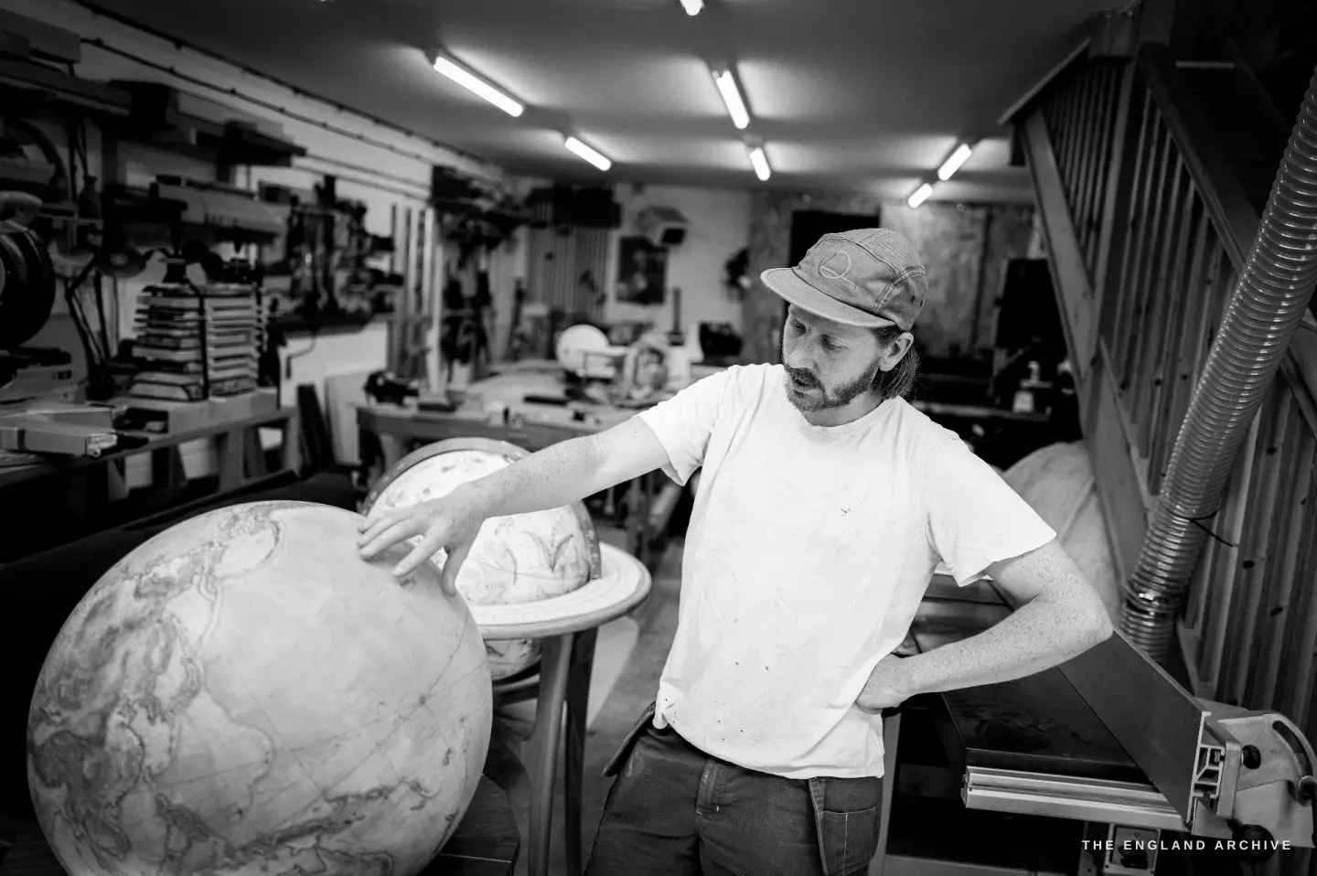 Louis standing among globes in his workshop, a hand resting on a large terrestrial globe, timber and tools behind.