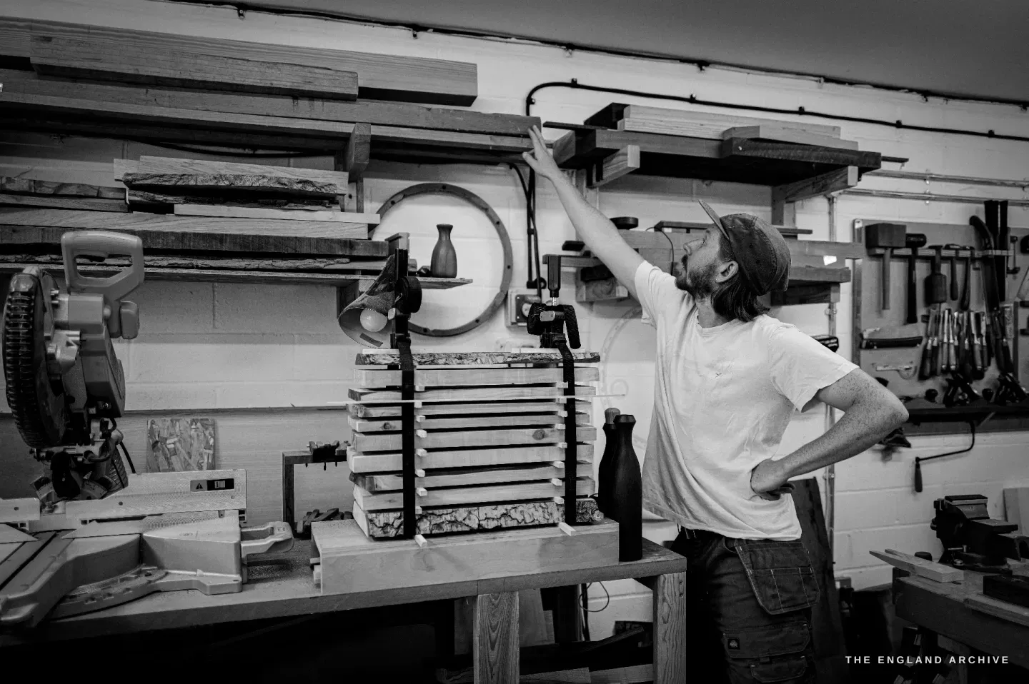 Louis reaching up to a timber rack at his bench, beside a clamped stack of laminated wood.