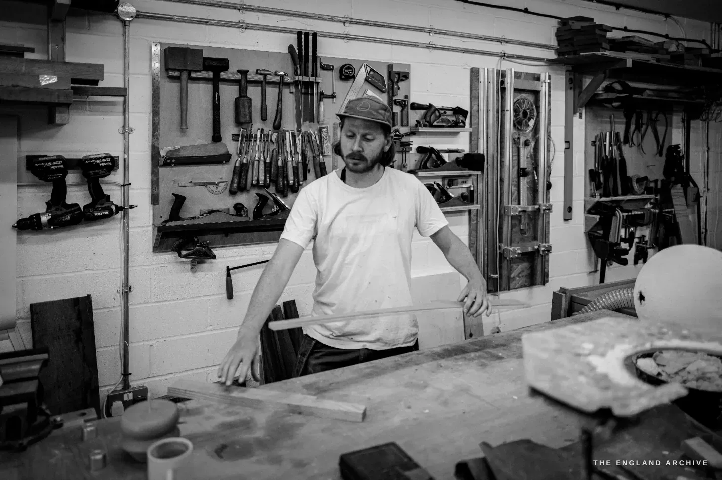 Louis at his workbench, hands on the bench, looking down at his work, a wall of chisels and mallets behind him.