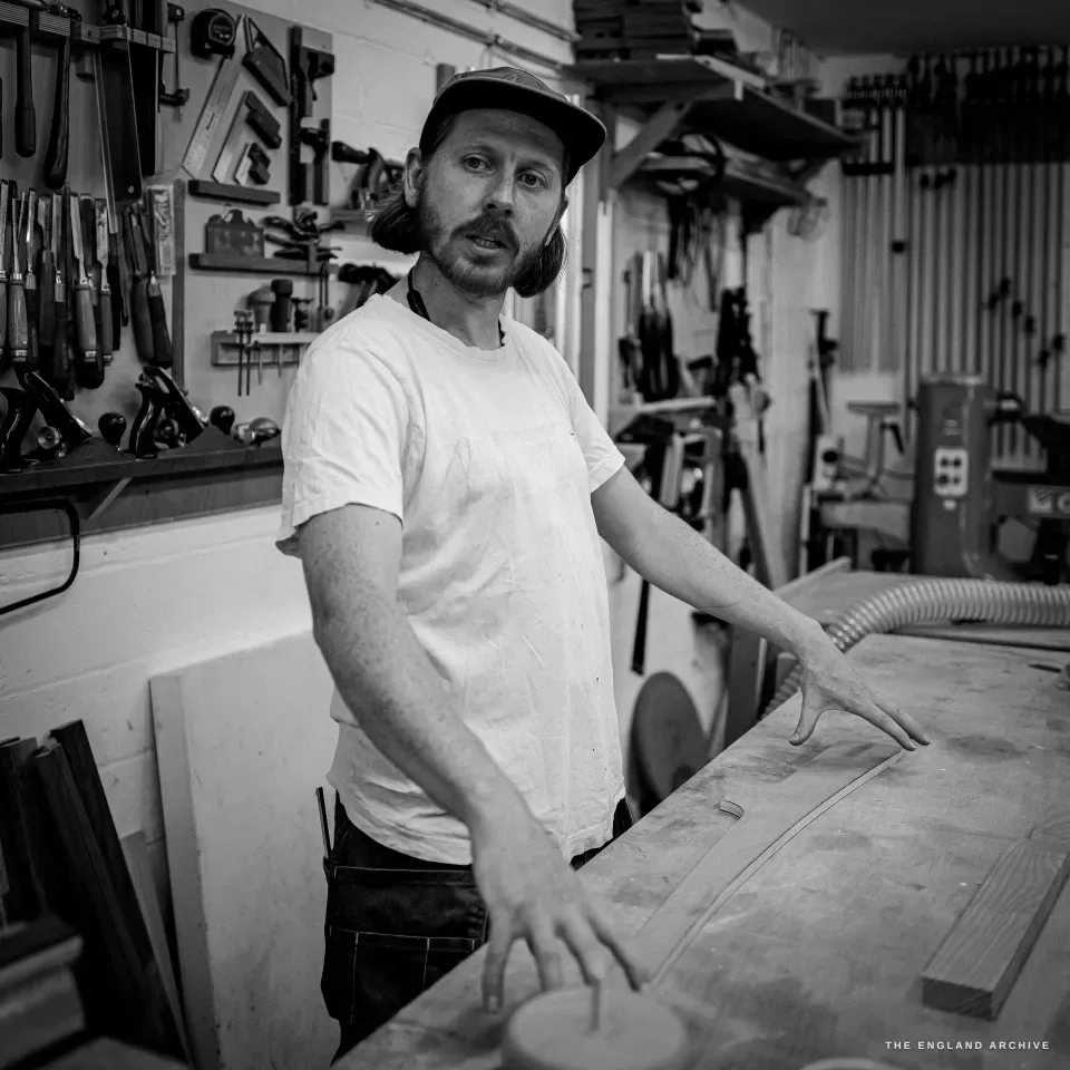 Louis Craig Carpenter at his workbench, looking to camera, woodworking tools racked on the wall behind him.