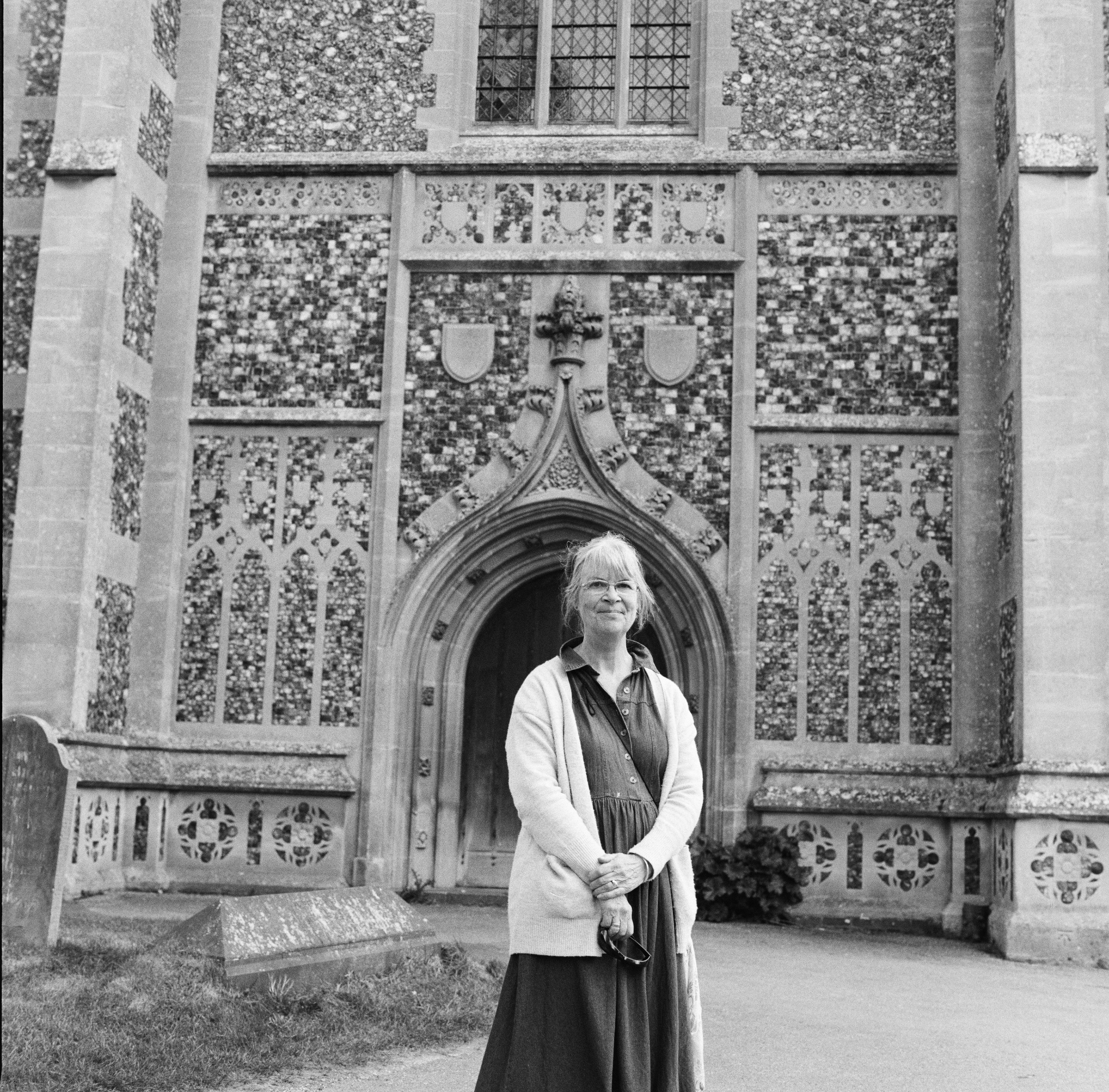 Melonie Clubb standing at the flushwork porch of Holy Trinity, Long Melford, hands clasped, without the booklet.