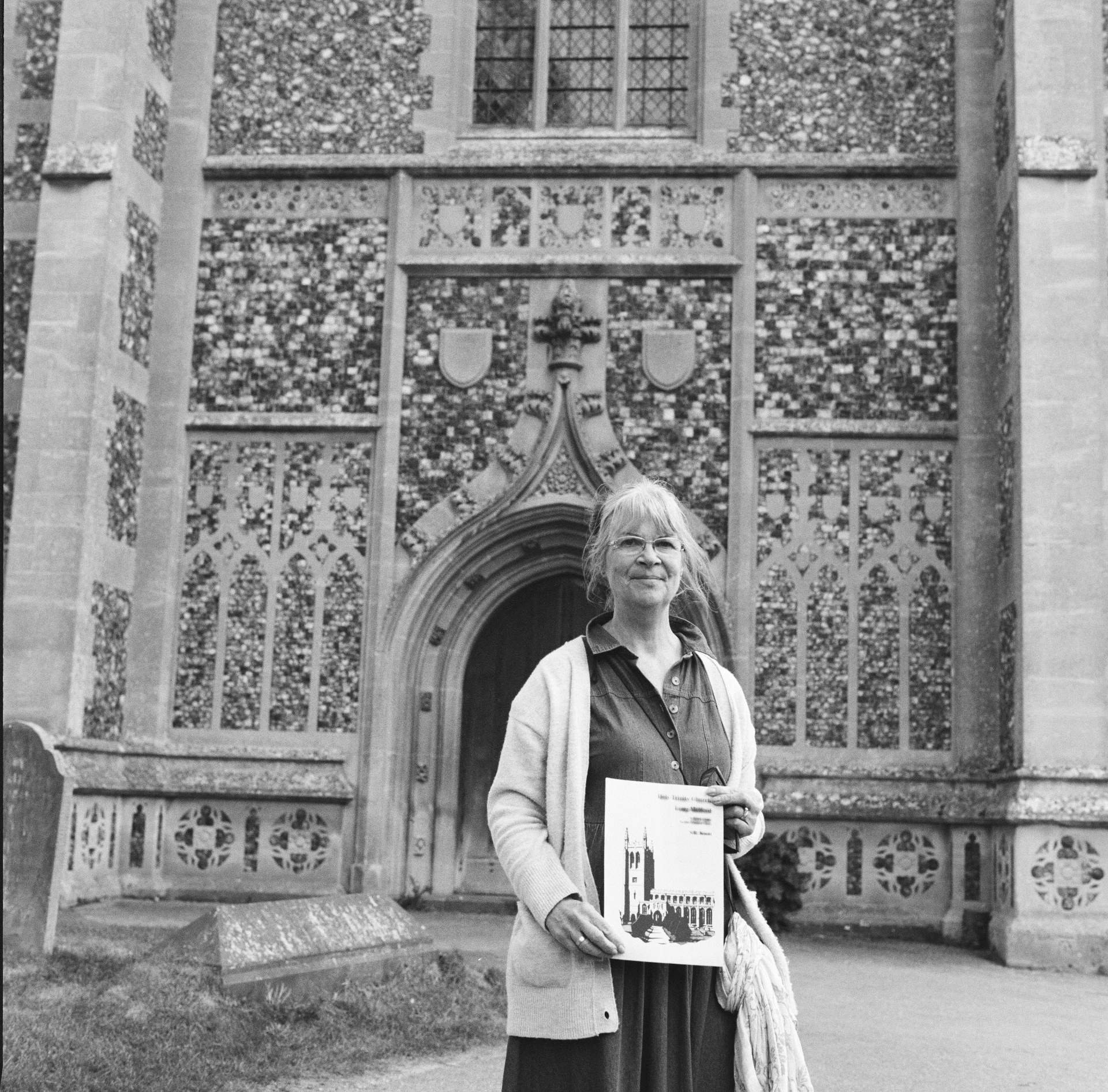Melonie Clubb standing at the flushwork porch of Holy Trinity, Long Melford, holding the Long Melford history booklet her father helped to produce.