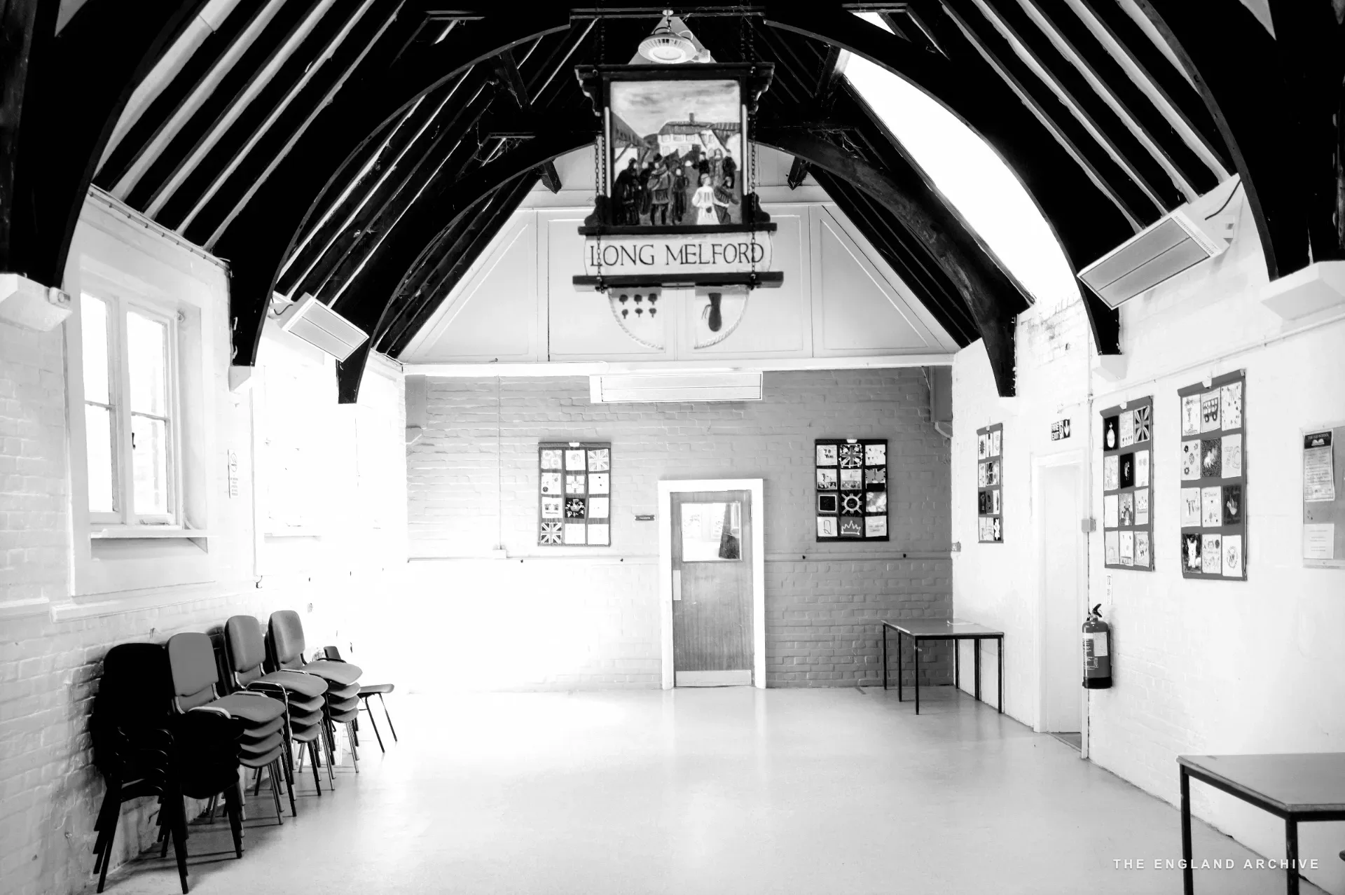 Community hall interior with exposed dark timber roof trusses and stacked red and blue chairs, a red wall at the far end.