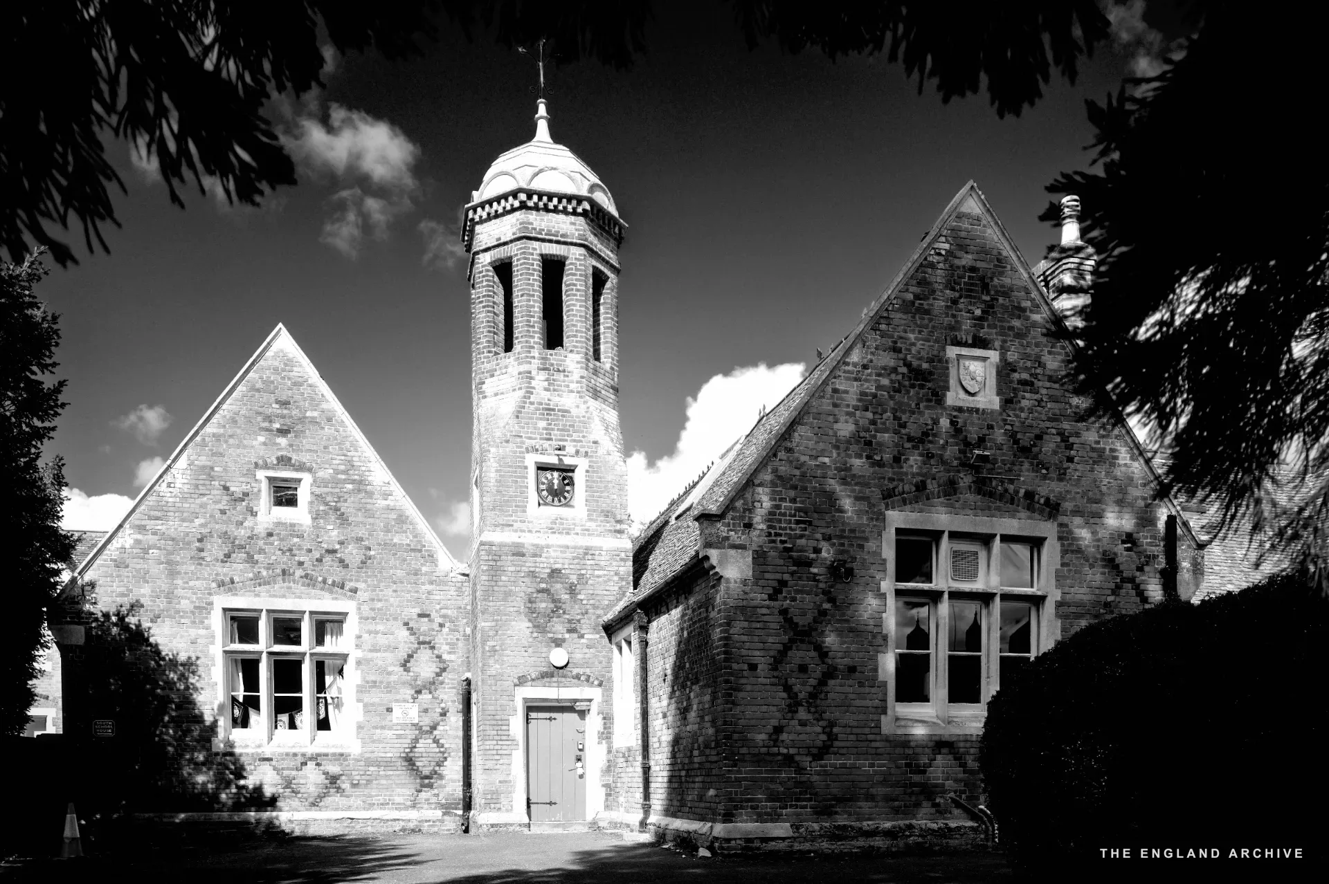 The Old School seen from the front garden, red brick with diaper brickwork and the central bell turret against a blue sky.