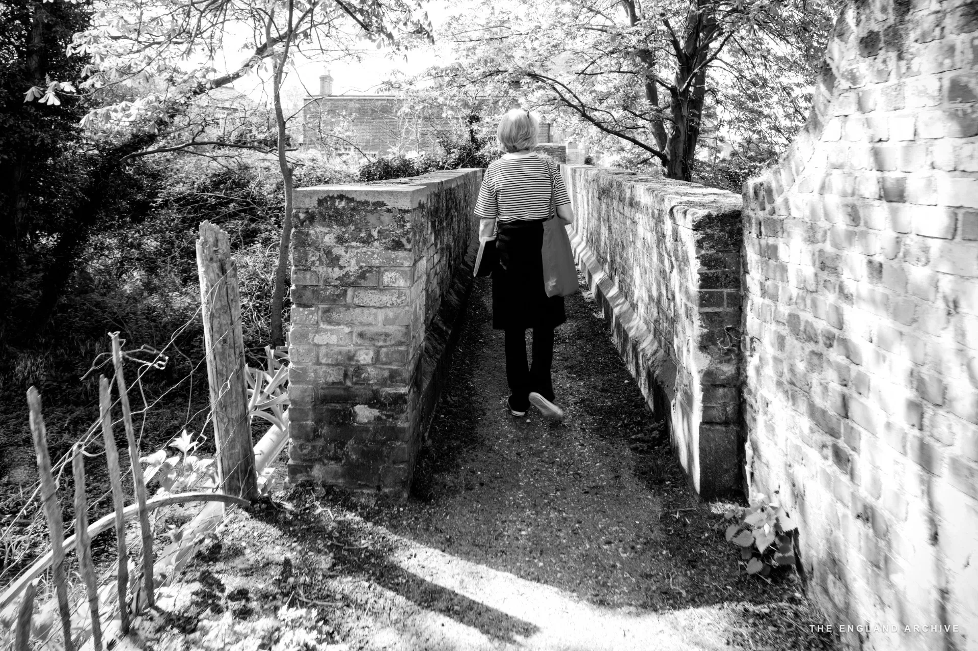 Julie Thomson walking away from the camera across a narrow brick footbridge with greenery on either side.