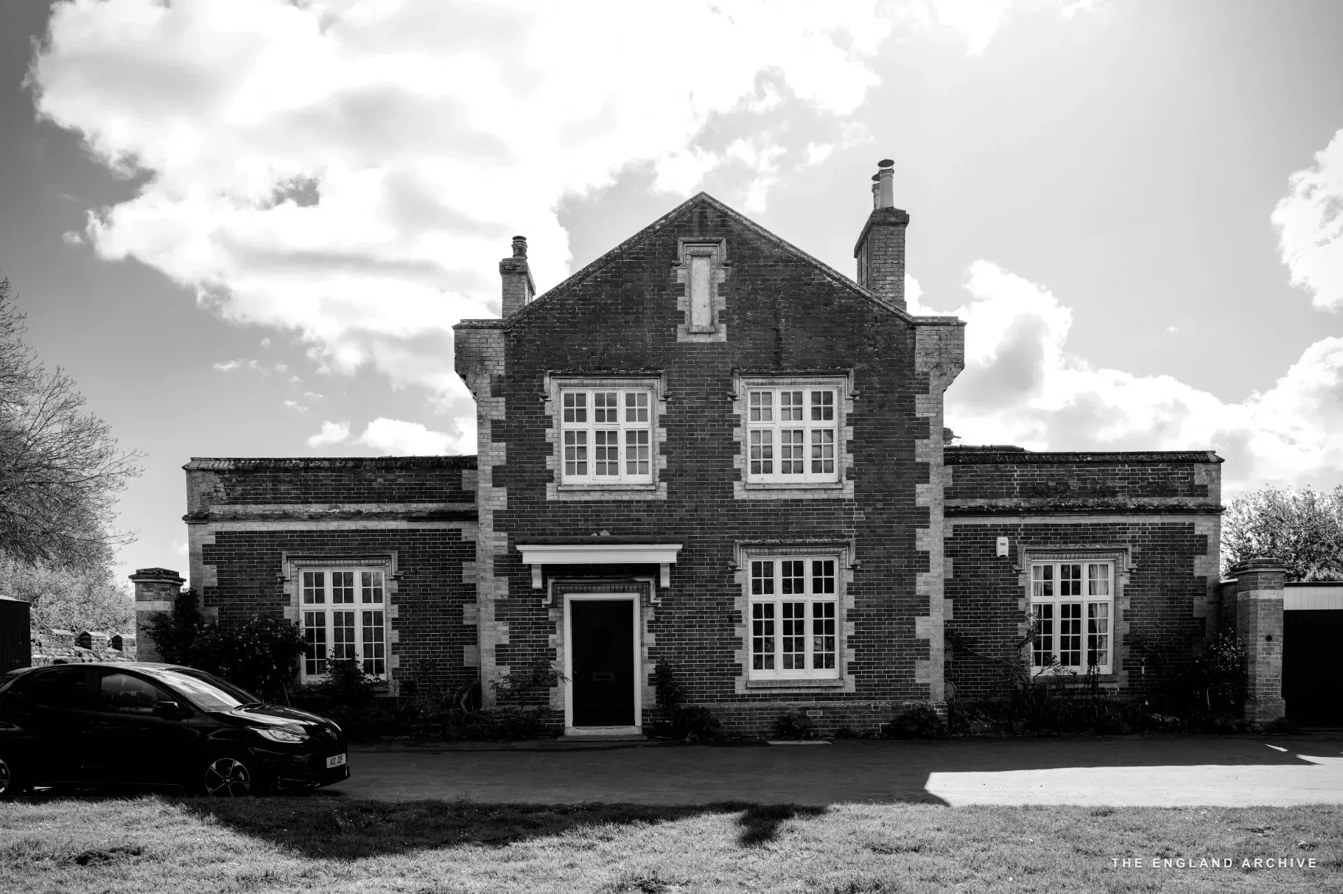 A symmetrical red-brick Victorian house with a blue door and a blue car parked on the lawn.