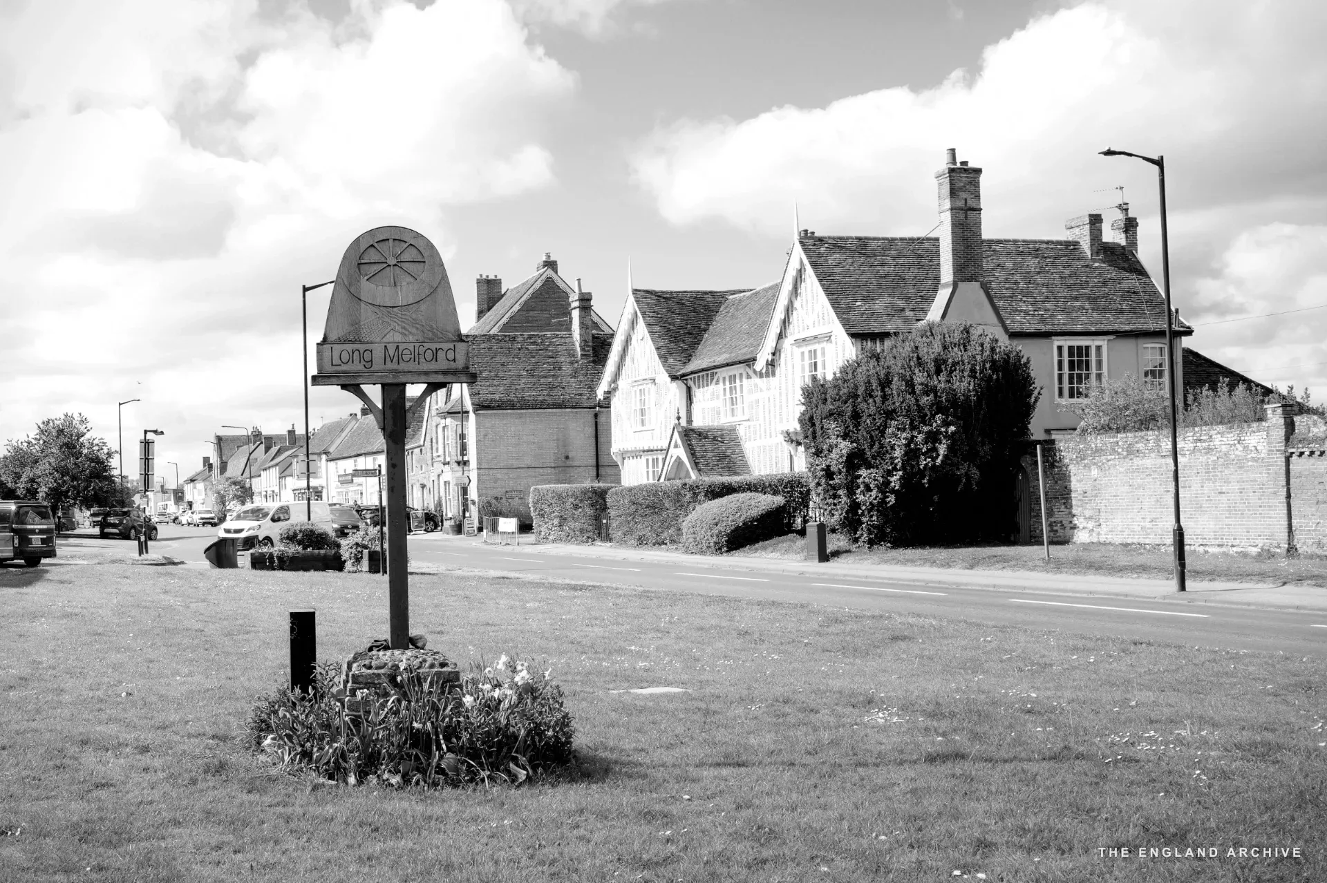 A 'Long Melford' village sign on a grass verge with a white Tudor timber-framed building behind.