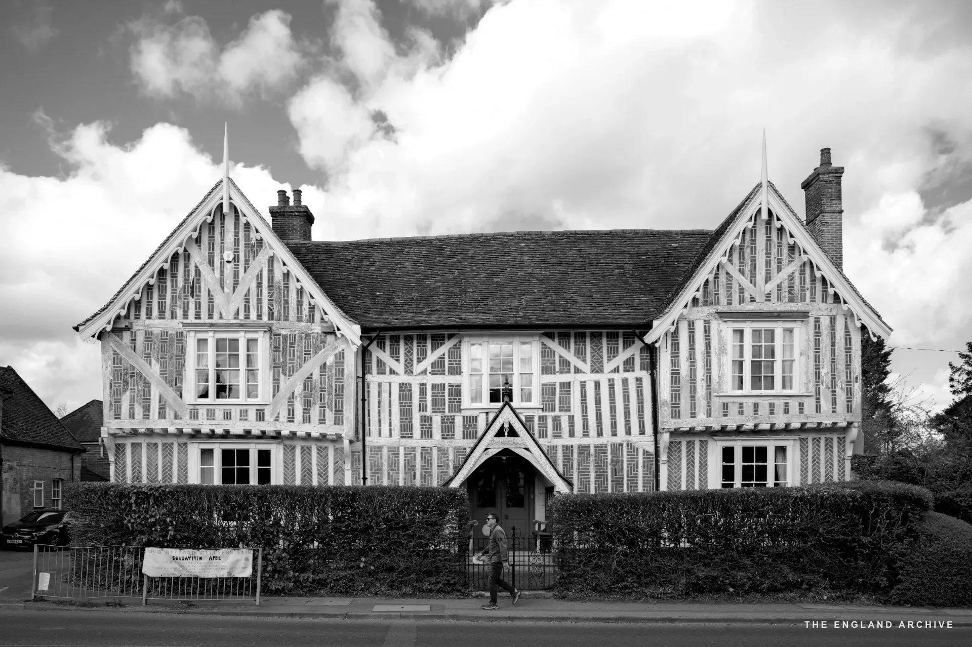 A large white and red brick Tudor house with decorative diaper patterning and two steep gables, a figure walking past on the pavement.