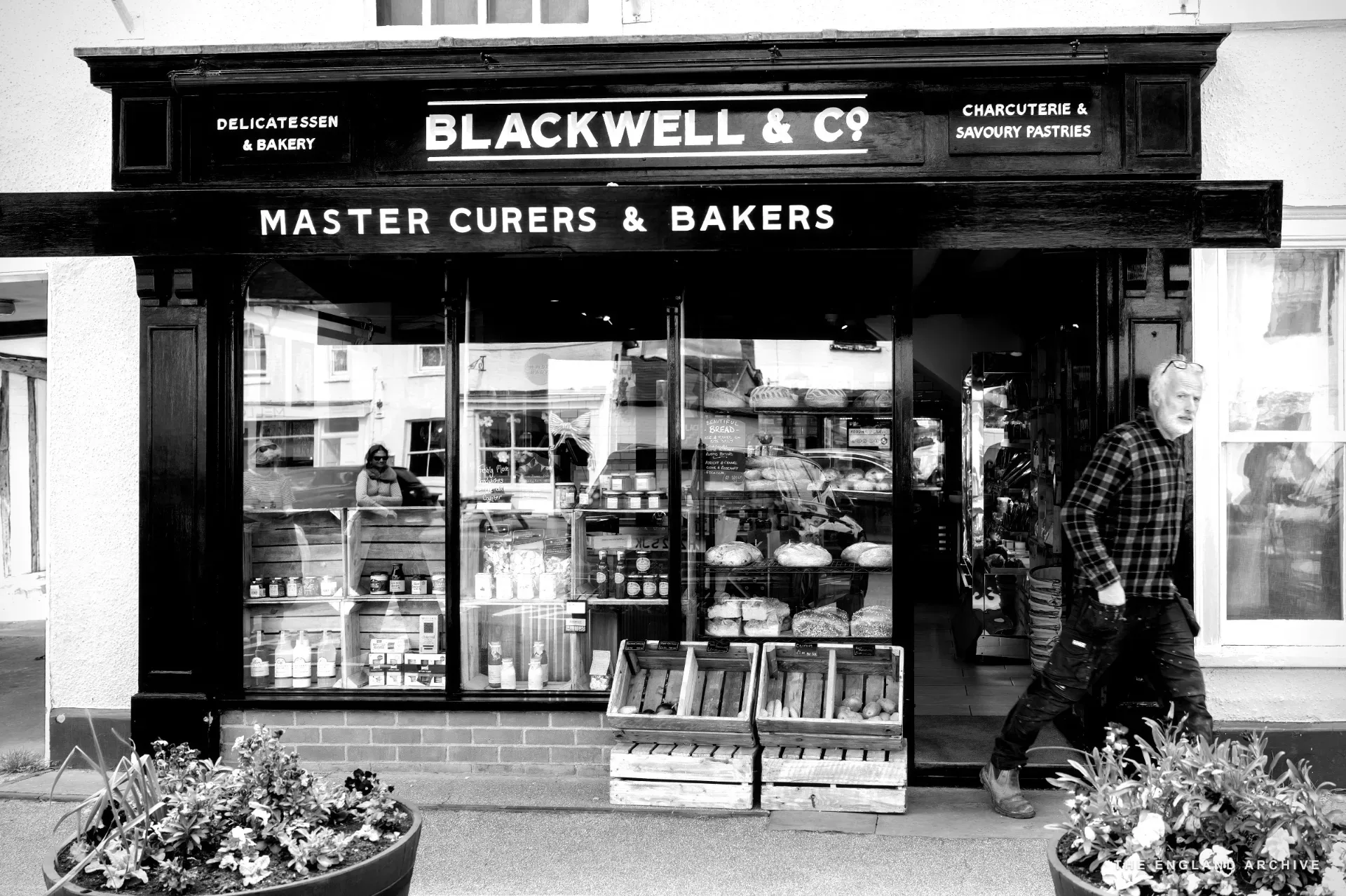 The 'Blackwell &amp; Co Master Curers &amp; Bakers' shop front with a pale pink wall, an A-frame chalkboard on the pavement and a wooden barrel planter.