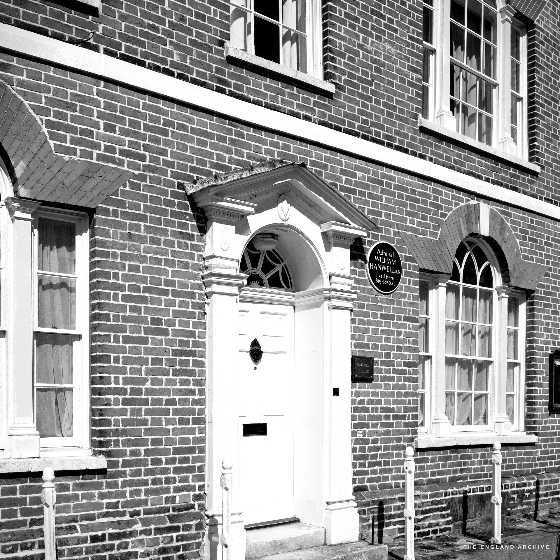 A cyclist in black and pink Lycra removing a helmet in the doorway of a cafe, a grey-haired woman in sunglasses walking past in the foreground.
