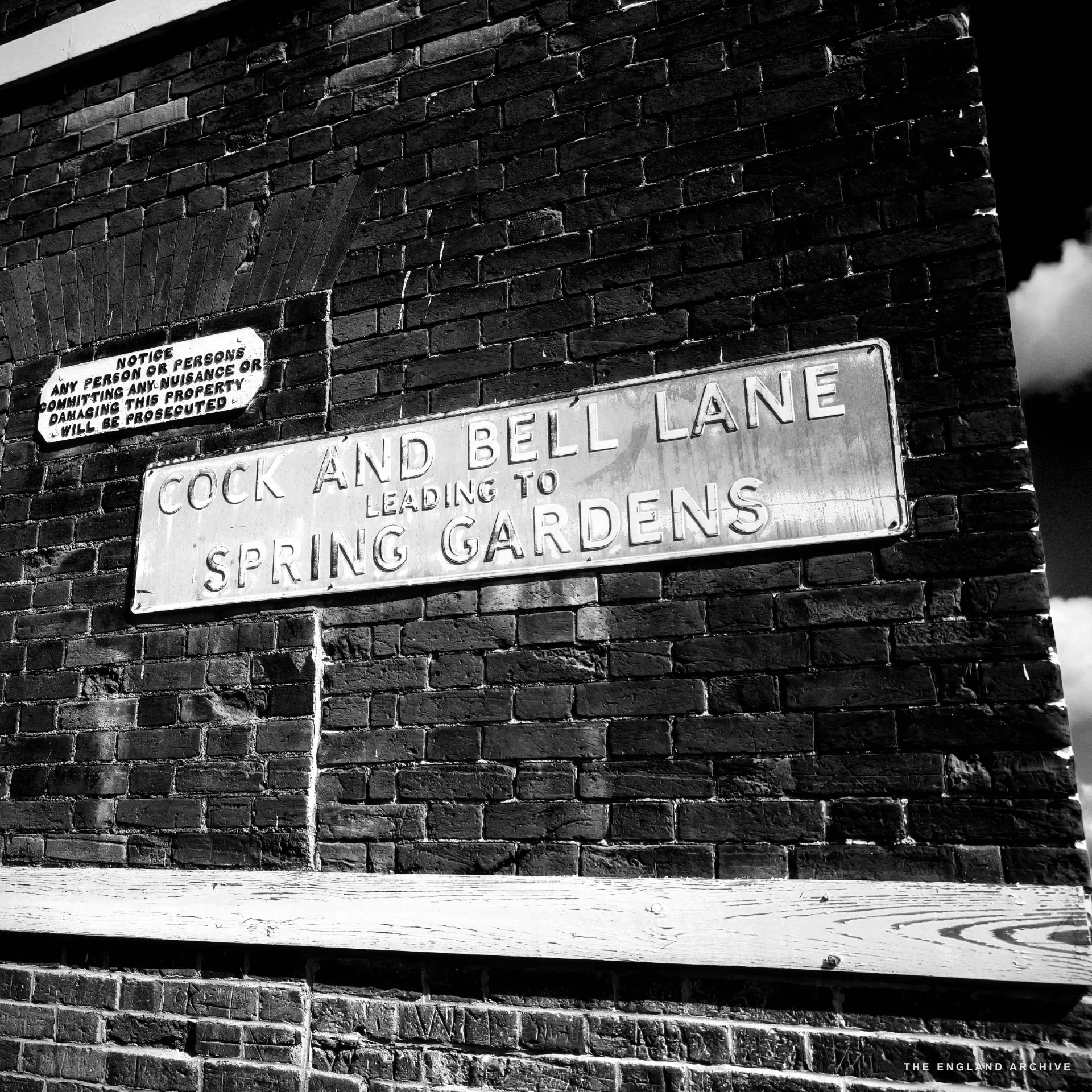 A black metal street sign reading 'COCK AND BELL LANE leading to SPRING GARDENS' fixed to a red brick wall, with a smaller prosecution notice above it.