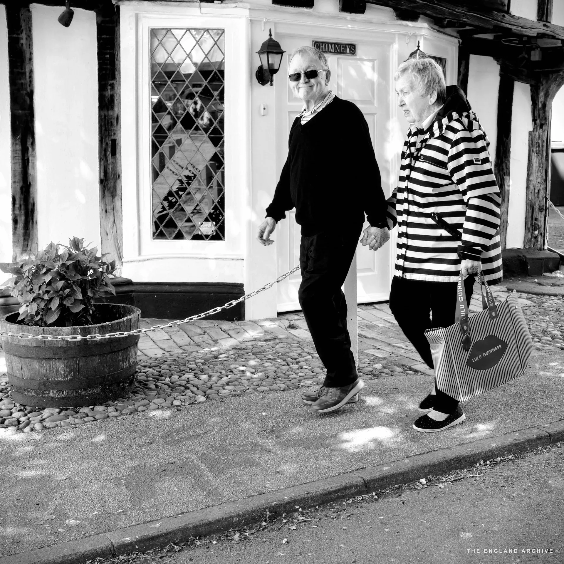 An older couple walking along a cobbled path past a leaded bay window marked 'CHIMNEYS', a red tote bag over the woman's shoulder.