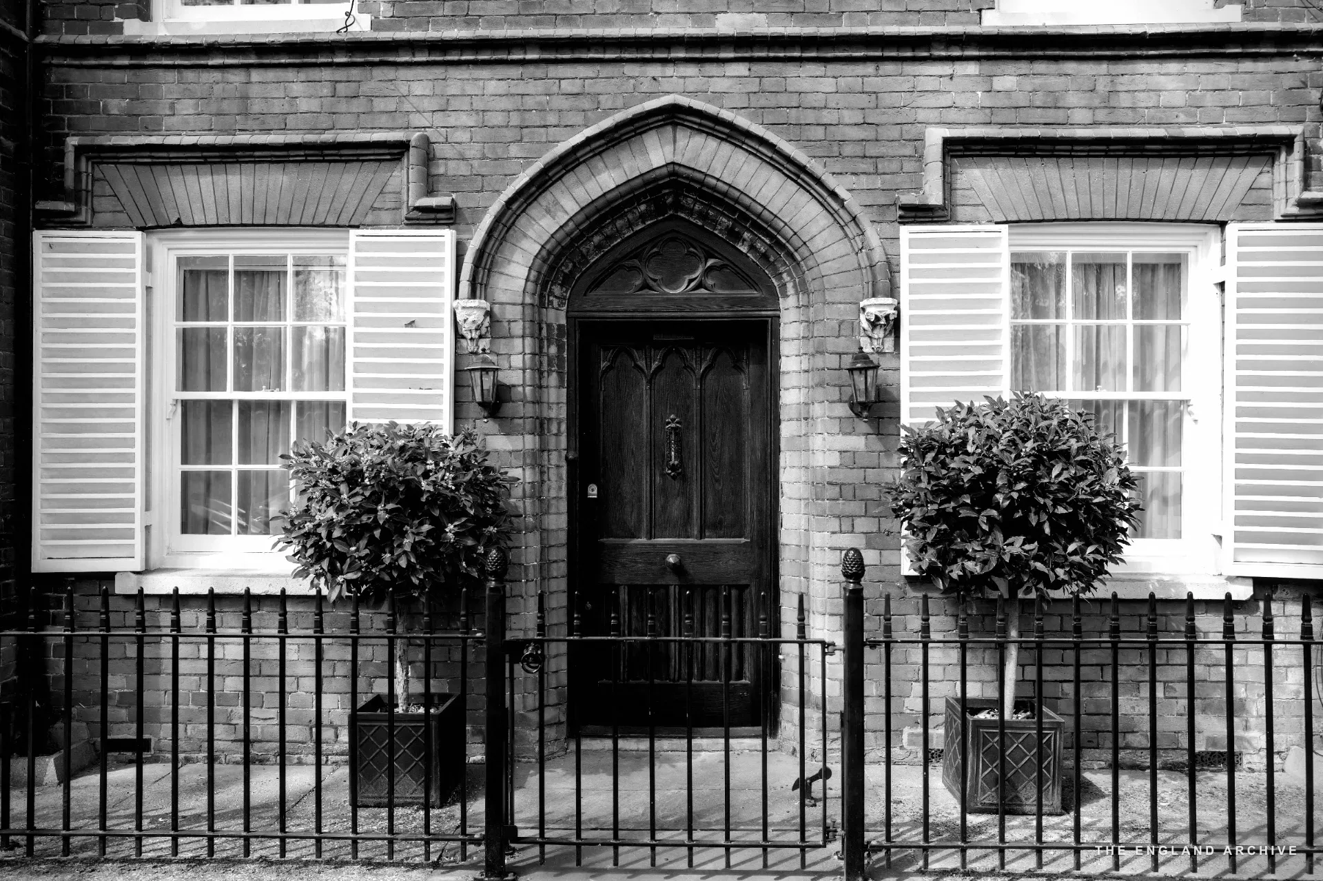 A red brick facade with an ogee-arched Gothic front door flanked by topiary bays in planters.