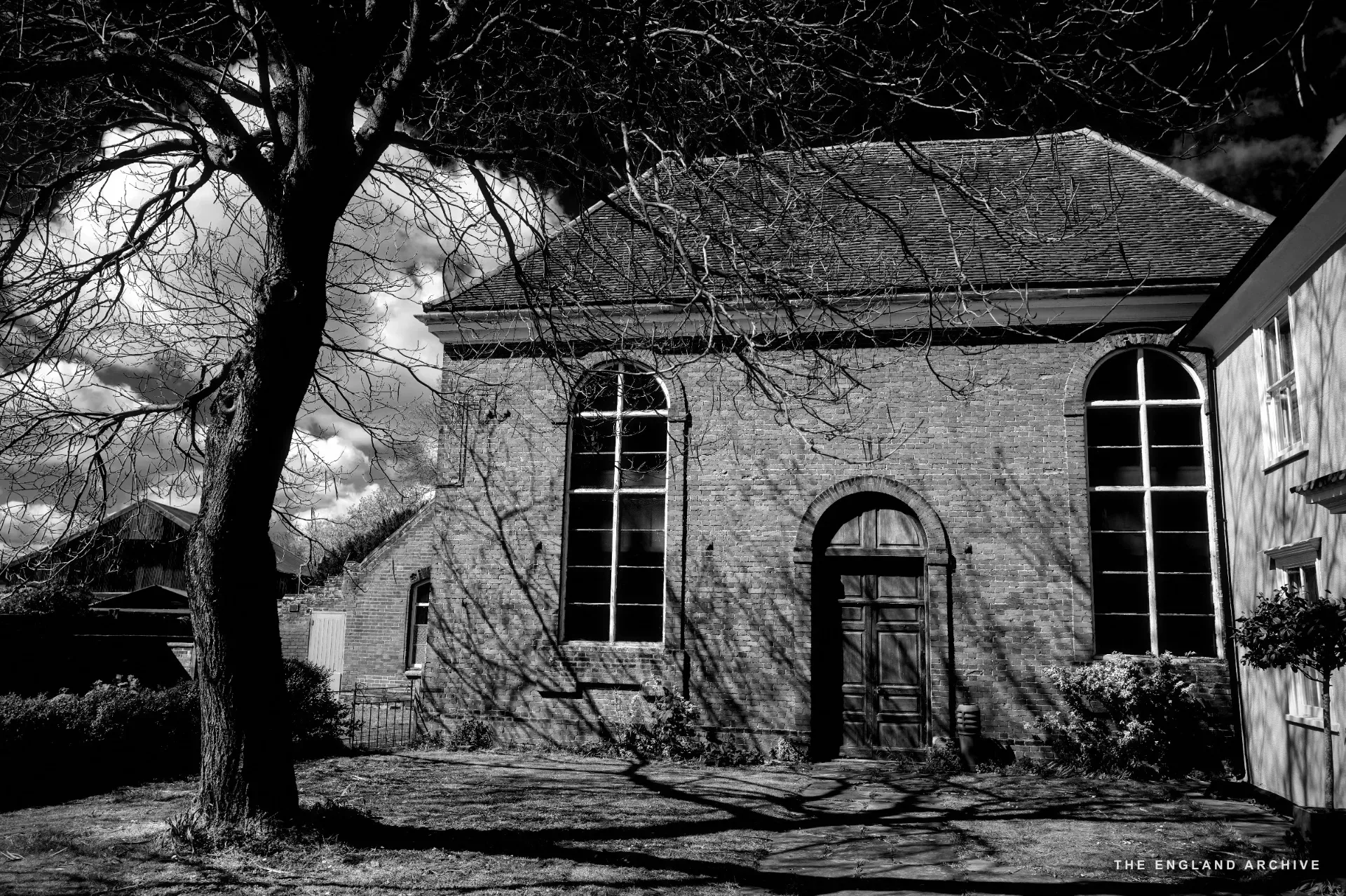 A red brick chapel with tall arched windows and a wooden door, set in a grass yard under a bare tree.