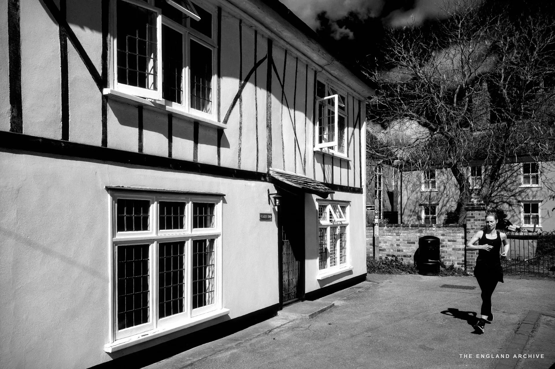 A white-rendered timber-framed cottage with leaded windows, a woman in black sportswear jogging past.