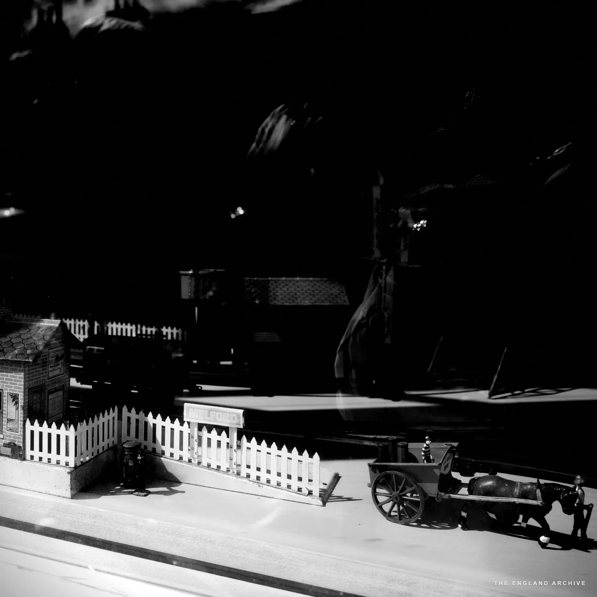 A shop window display of a model railway diorama with a 'MELFORD' station sign and a horse-drawn cart figure, street reflections in the glass.