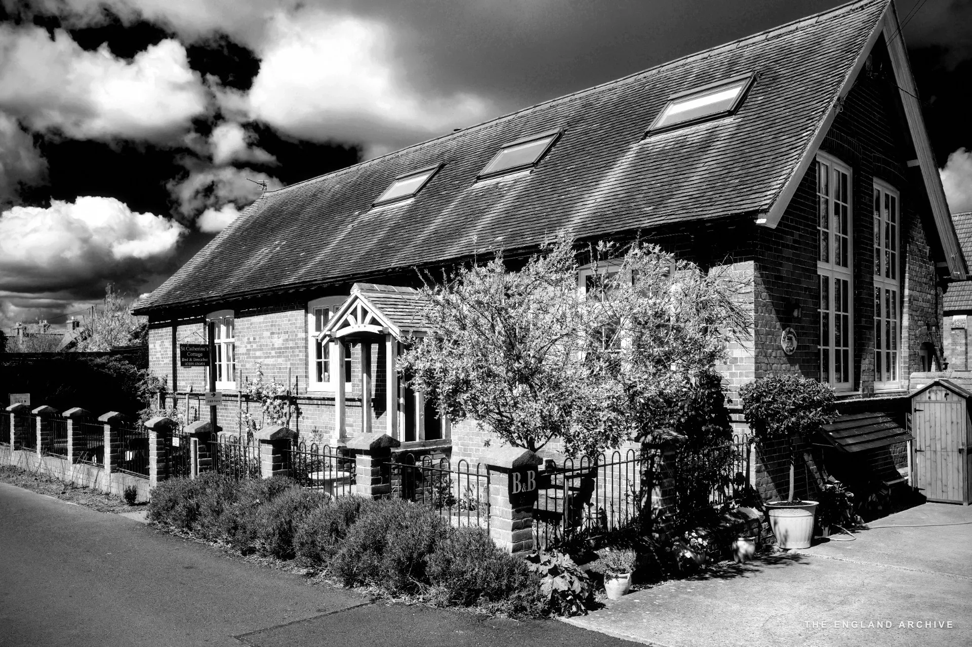 A brick-built converted cottage with a pitched roof, a small 'B&amp;B' sign on the gate and lavender bushes along the low wall.