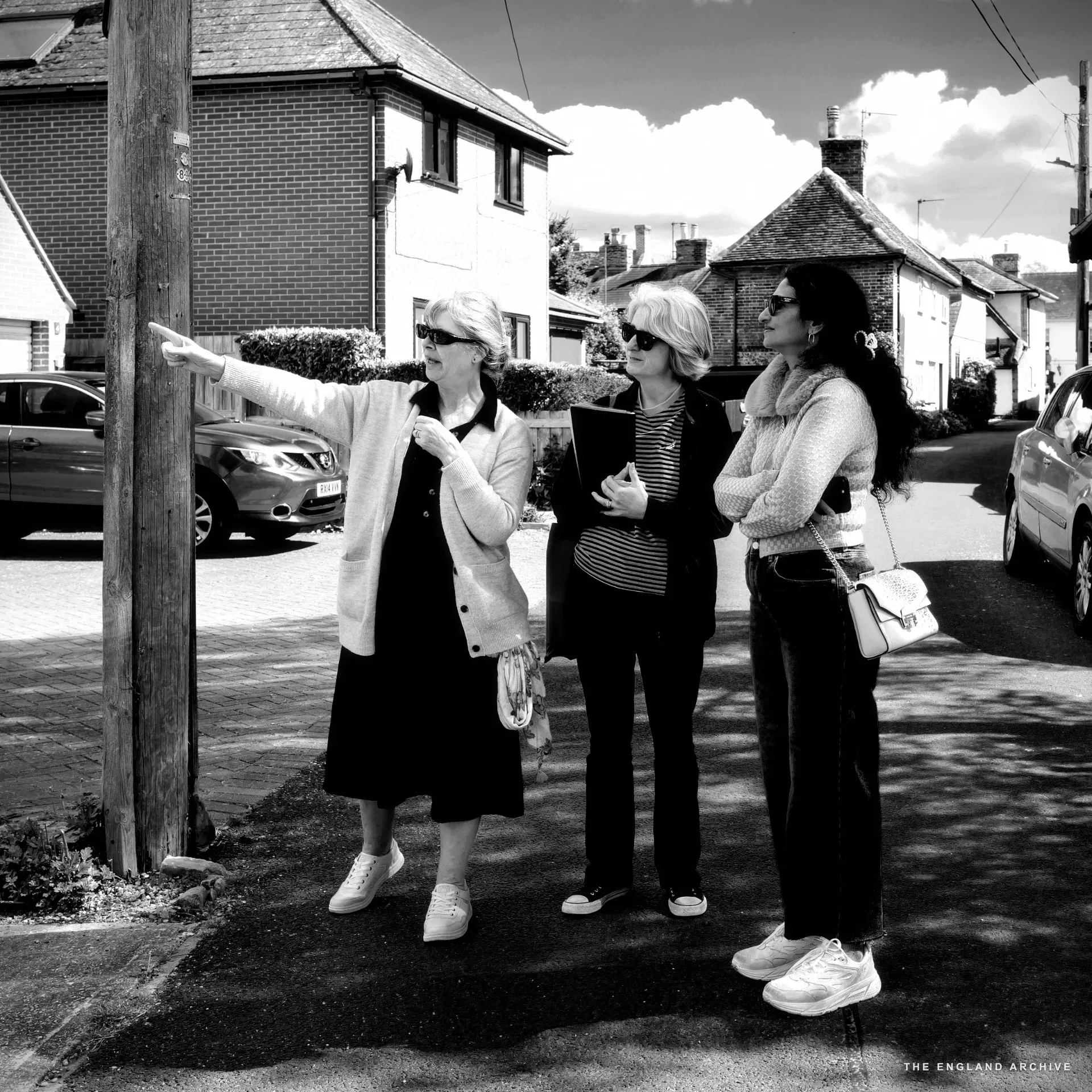 Three women on a residential street: Julie pointing at a house, Melonie with a blue folder and red scarf, Bhavani in a pink checked top.