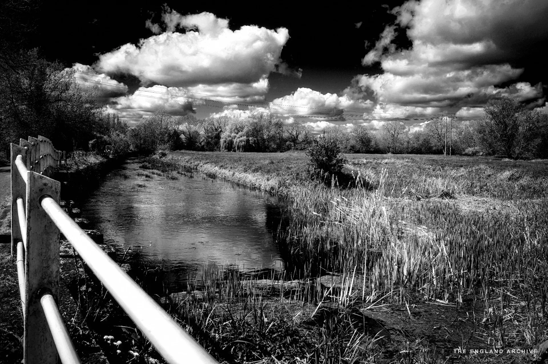 A small river with a white post-and-rail fence on the left, reed beds along the bank, open fields and a big cumulus sky beyond.