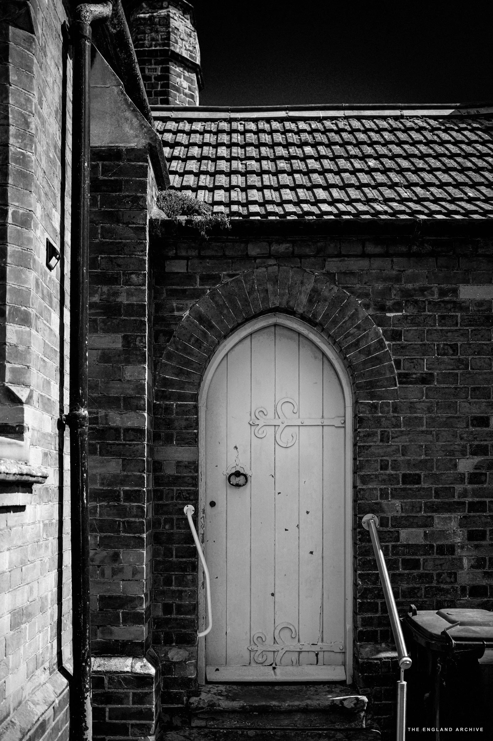 A narrow arched white-painted wooden door set in red brick, a pantile roof above.