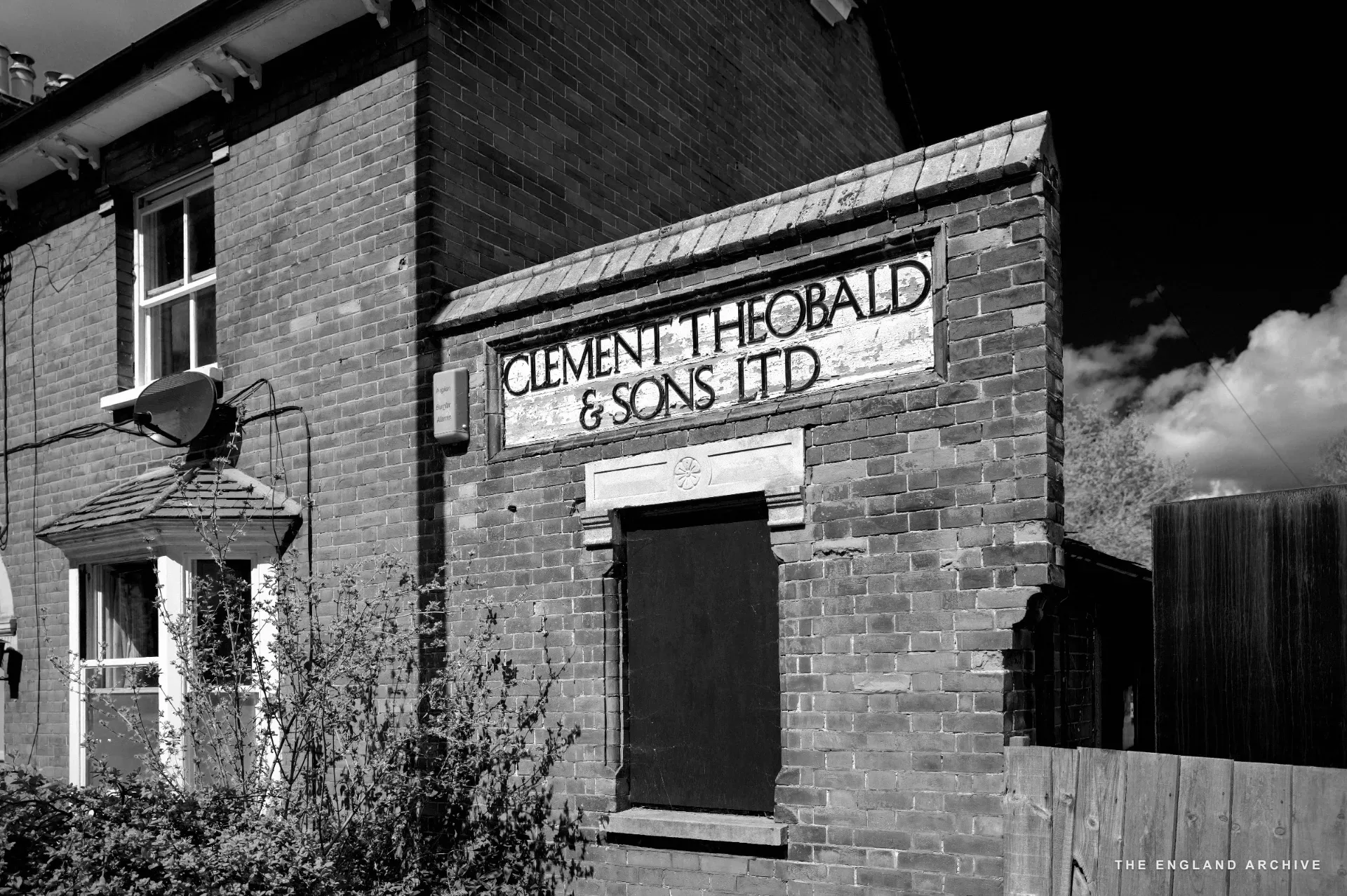 A red brick warehouse gable end with a painted sign reading 'CLEMENT THEOBALD &amp; SONS LTD' above a boarded window.