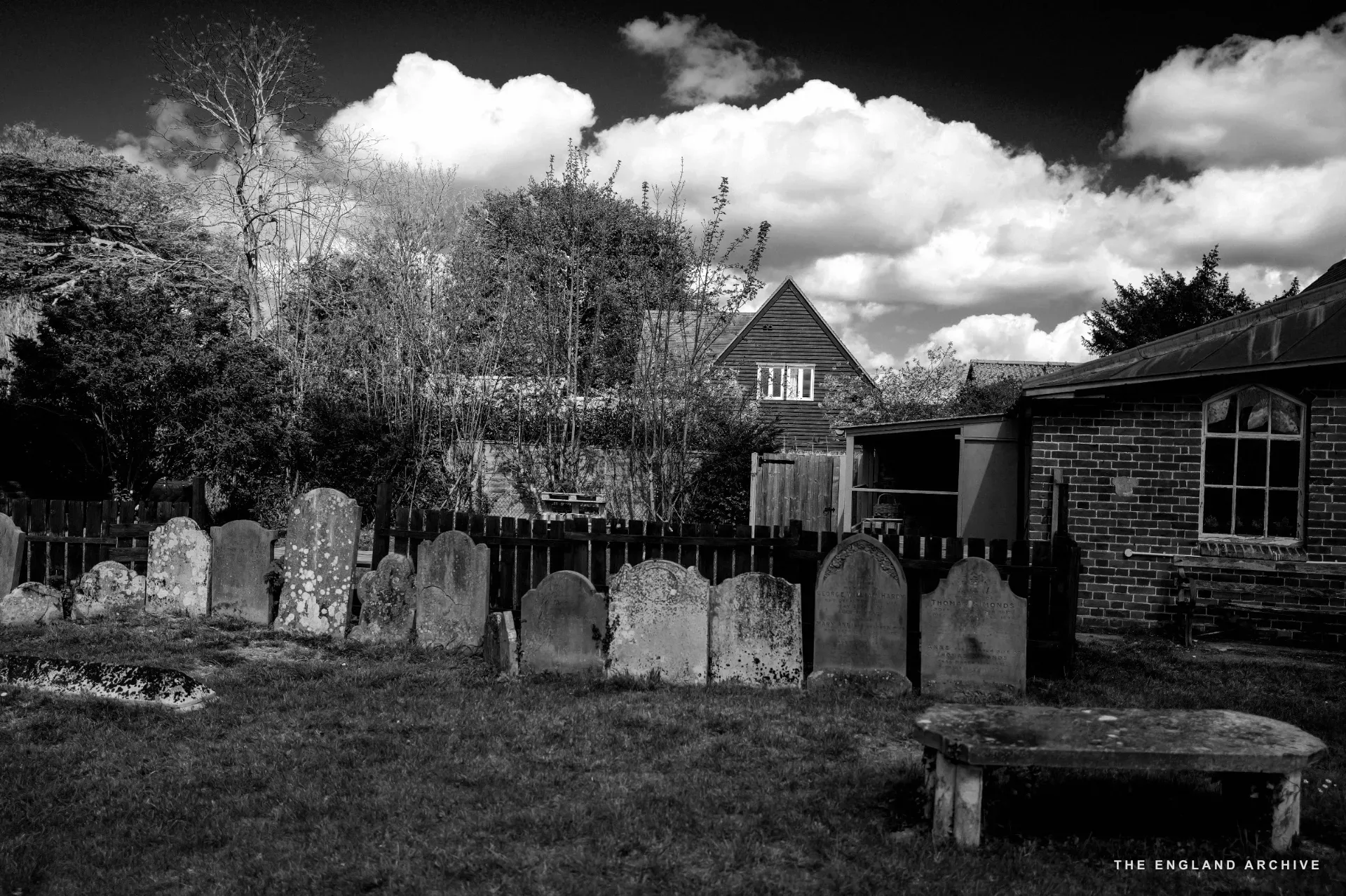 A row of weathered gravestones standing against a fenced green yard, a brick chapel with clerestory windows to the right.