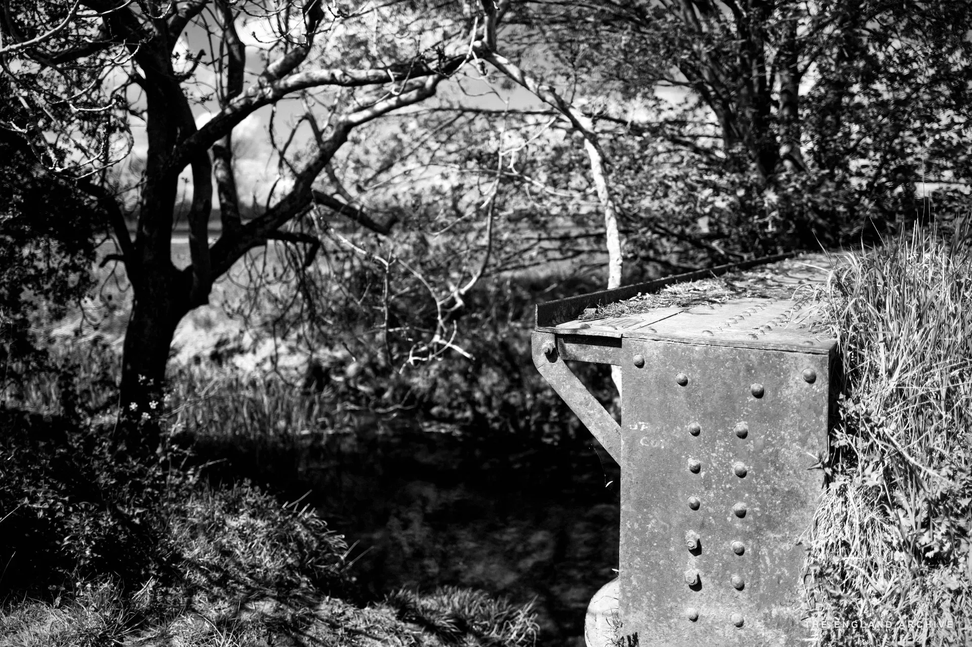 A side view of the same rusted iron girder bridge from the stream bank, looking out across open fields.