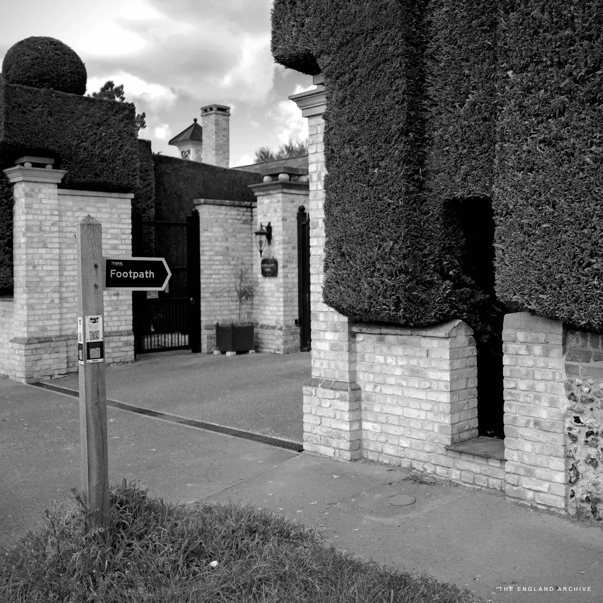 Tall clipped yew hedges flanking a gated drive with a black 'Footpath' signpost and brick gate piers.