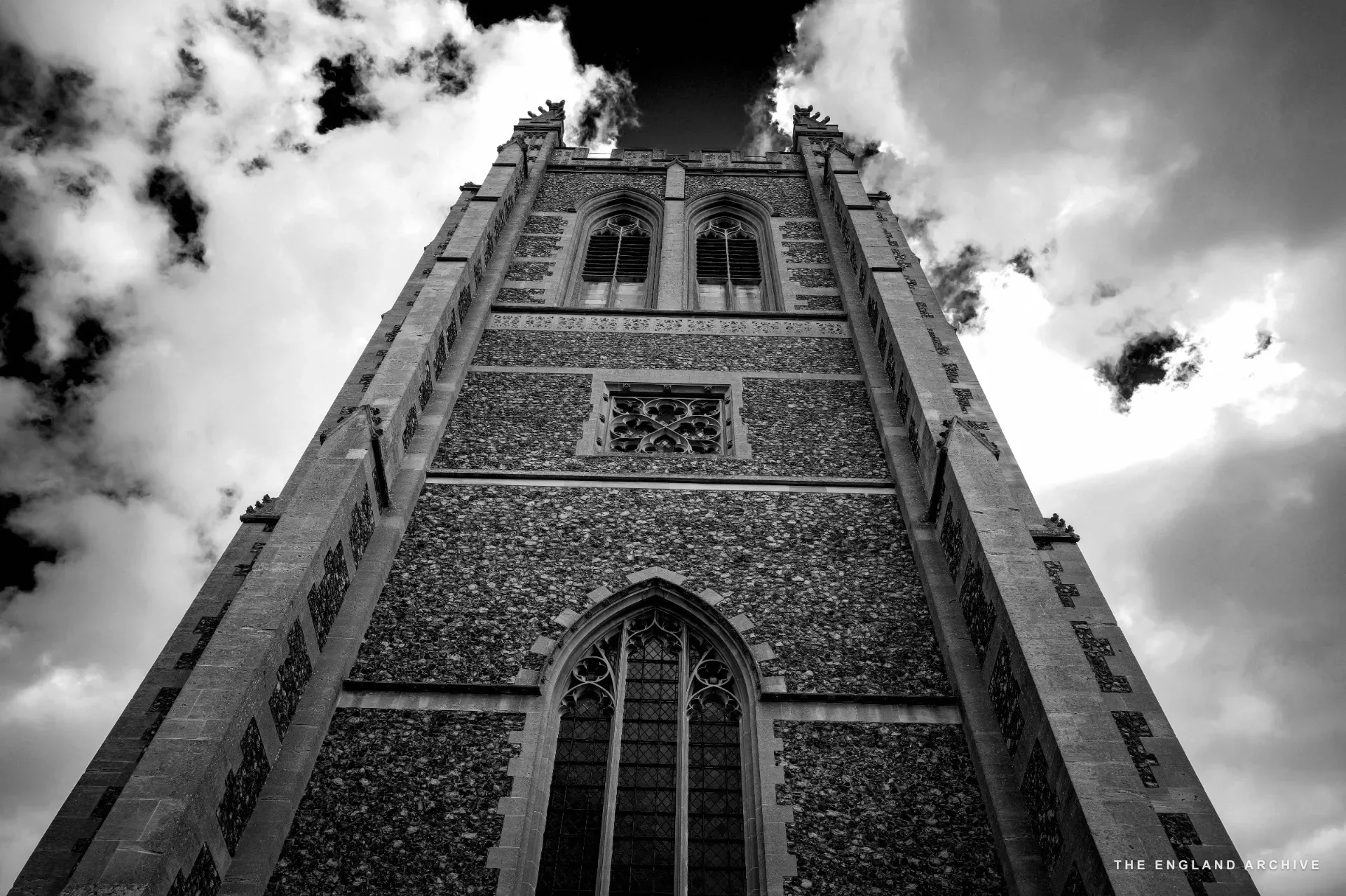 An upward view of Holy Trinity's tower, its flint and stone chequerboard flushwork catching the light, against a blue sky with clouds.