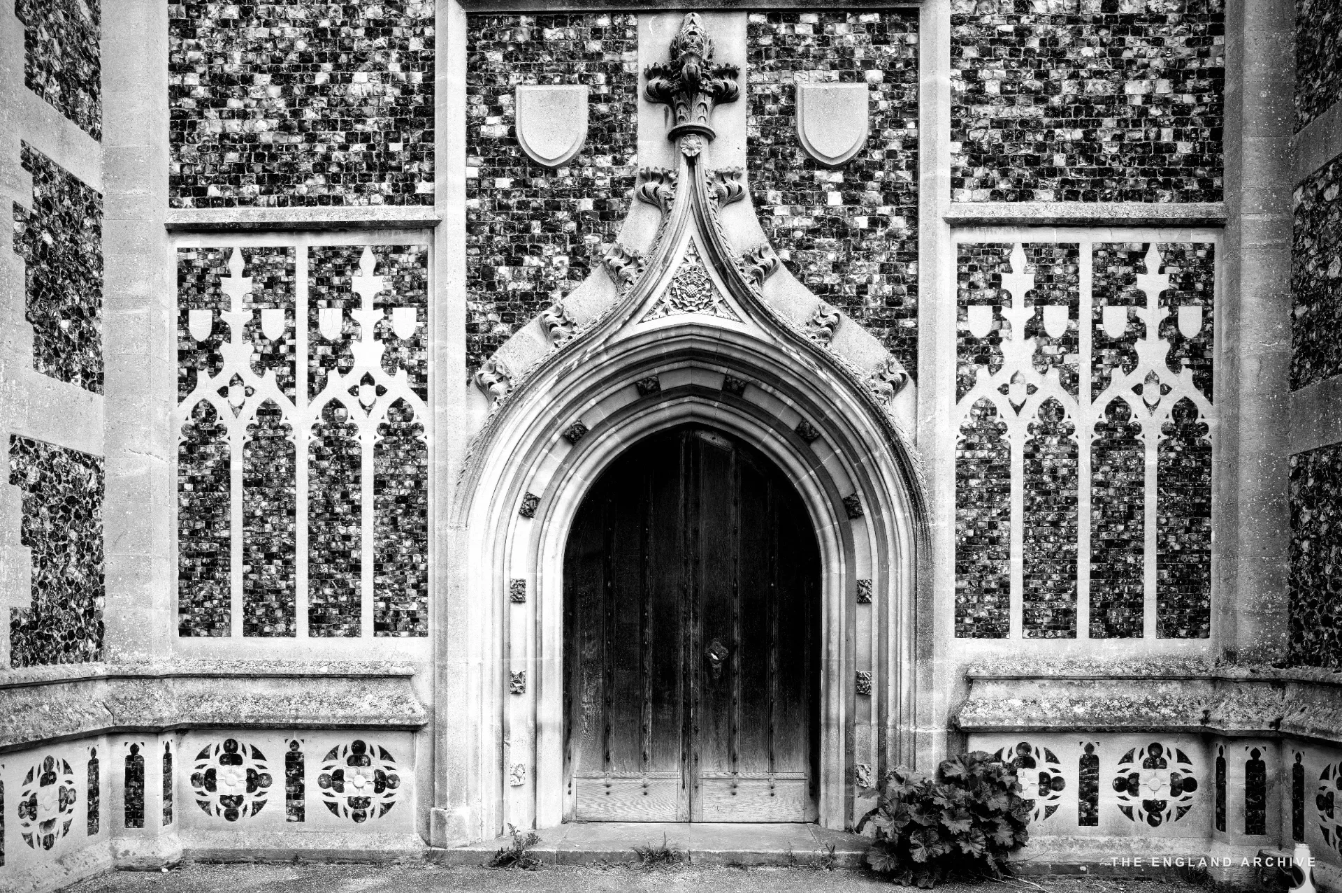 The arched Perpendicular doorway of Holy Trinity, flushwork flint and stone panels either side, a plant in the corner.
