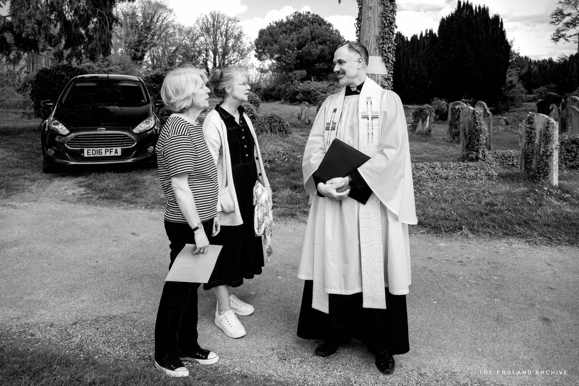 The vicar in a white surplice and black cassock standing in the churchyard talking with Julie Thomson and Melonie Clubb.