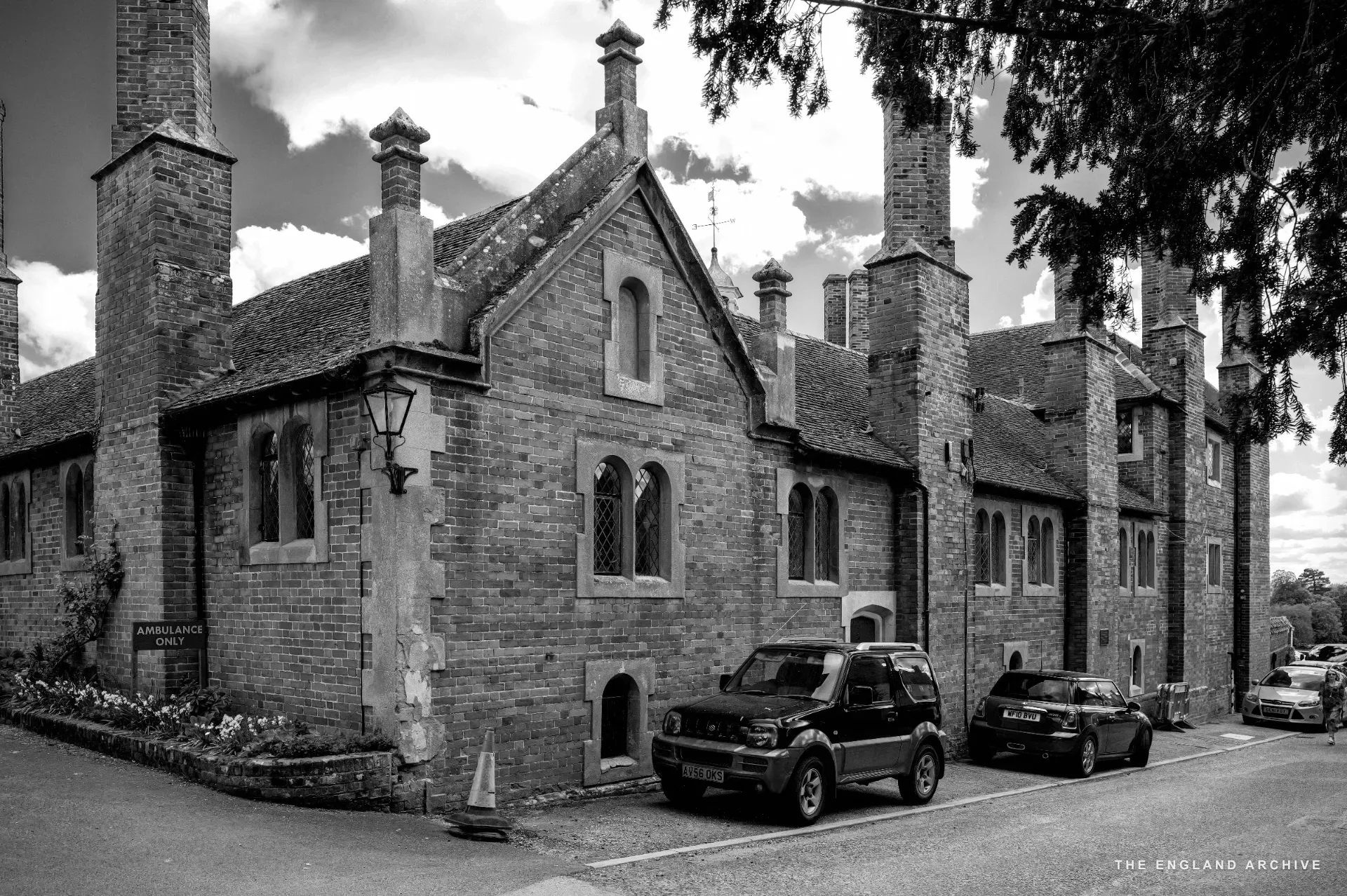The Tudor red-brick almshouse range of Trinity Hospital with tall chimneys and Gothic windows, cars parked at the side elevation.